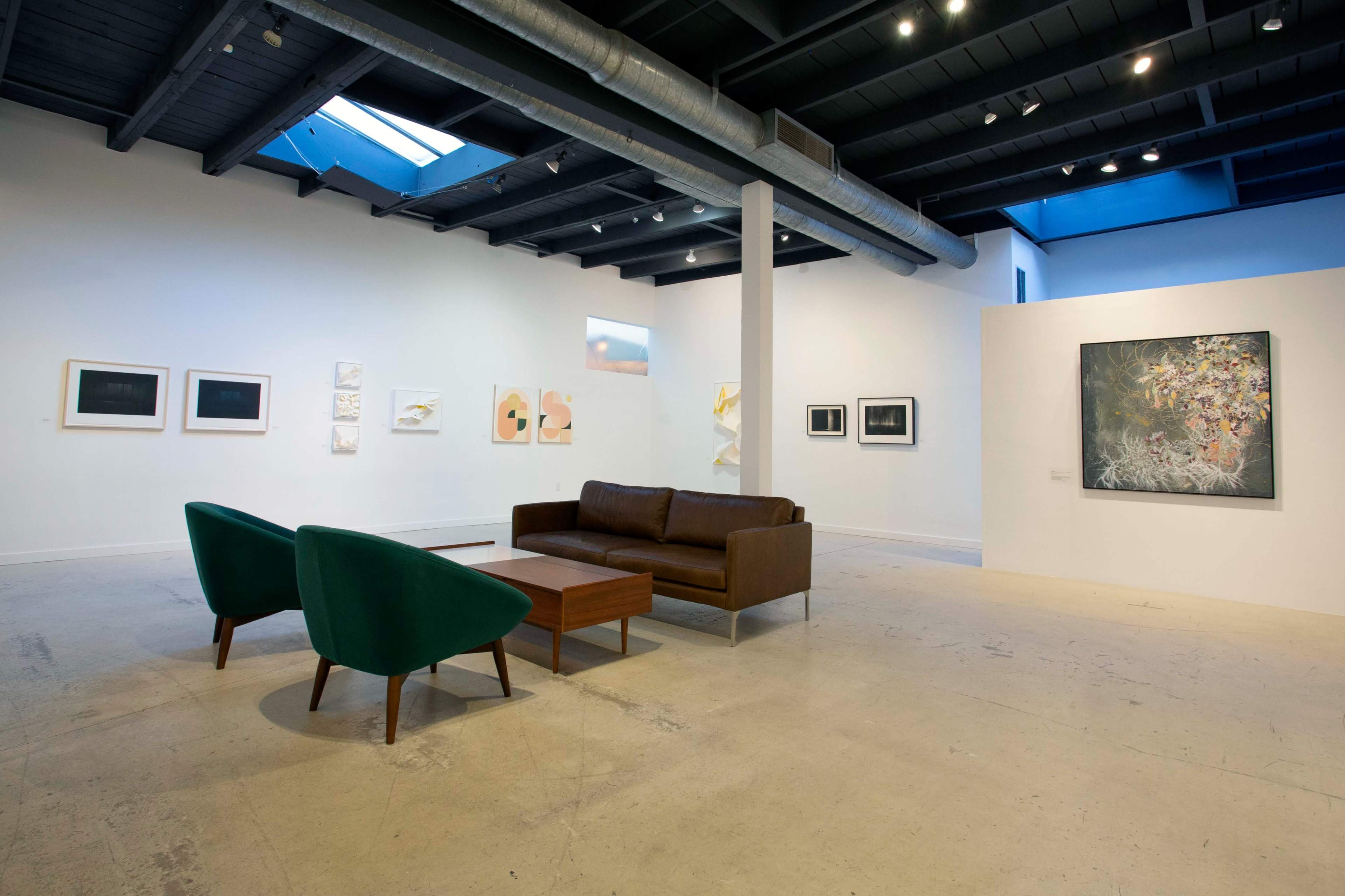 A contemporary art gallery featuring a brown sofa and two green chairs in the foreground, with various framed artworks displayed on the walls.