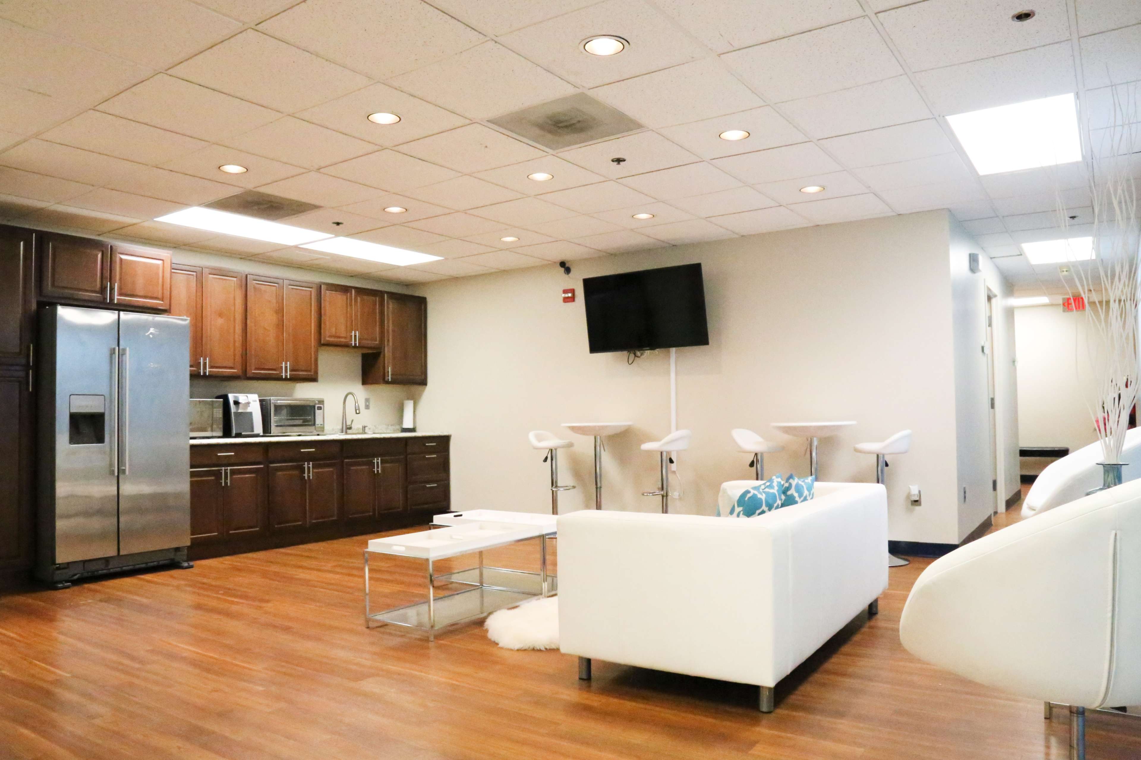 A modern lounge area with a kitchen, including stainless steel appliances, white furniture, and a wall-mounted television.