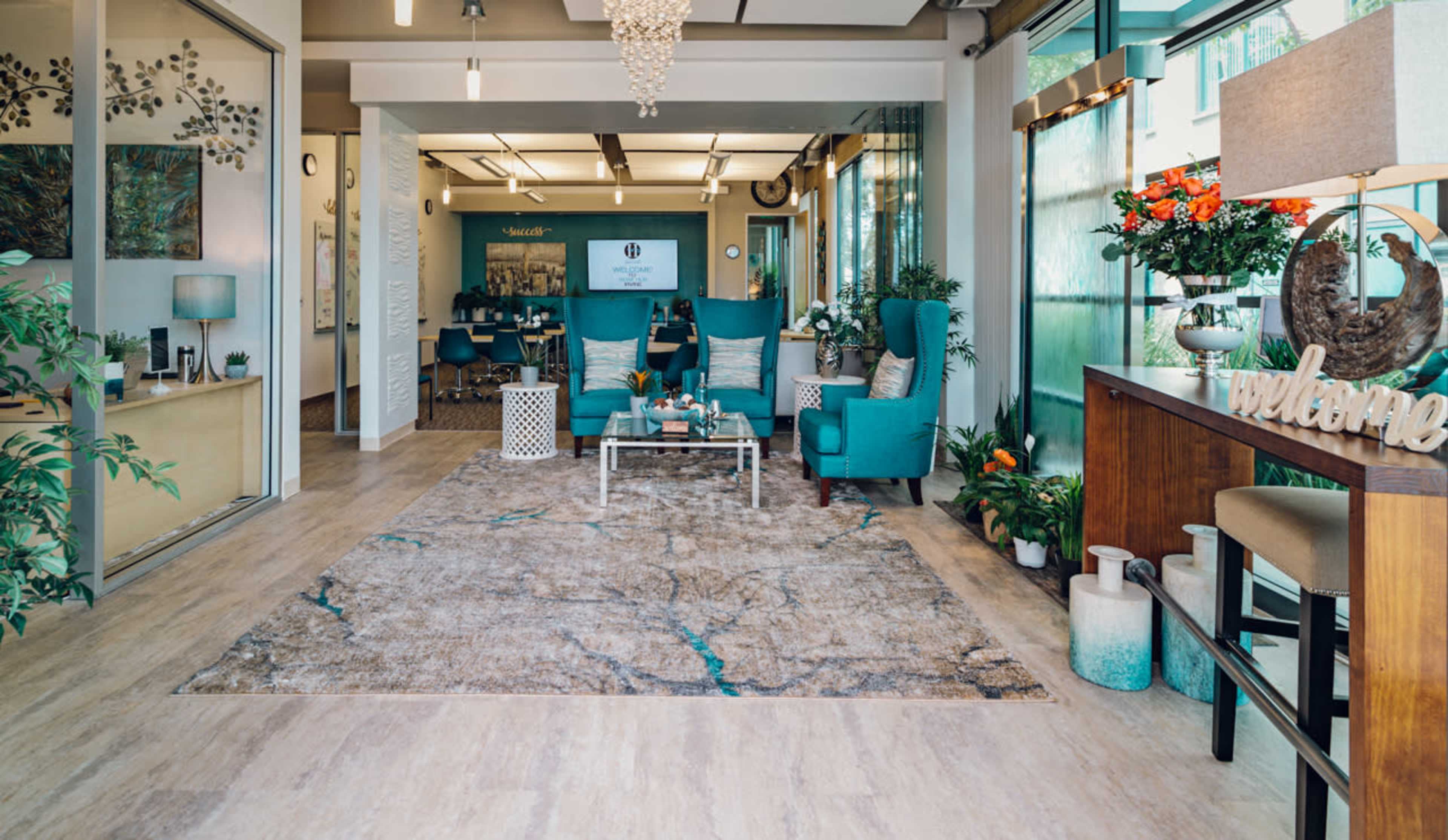 A modern reception area with teal seating, a decorative rug, and potted plants.
