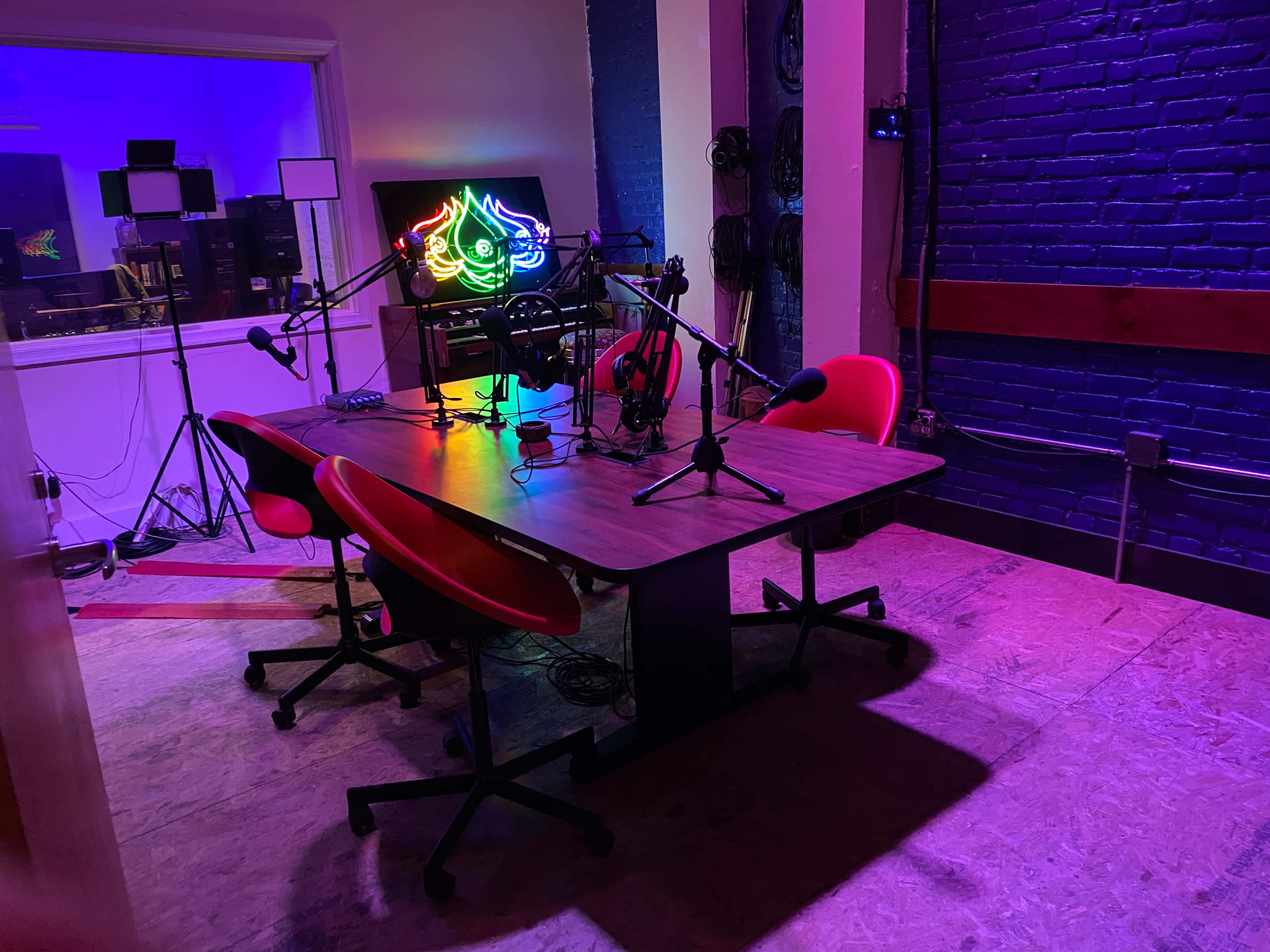 A podcast studio features a table with two red chairs, microphones, and colorful lighting against a blue-brick wall.