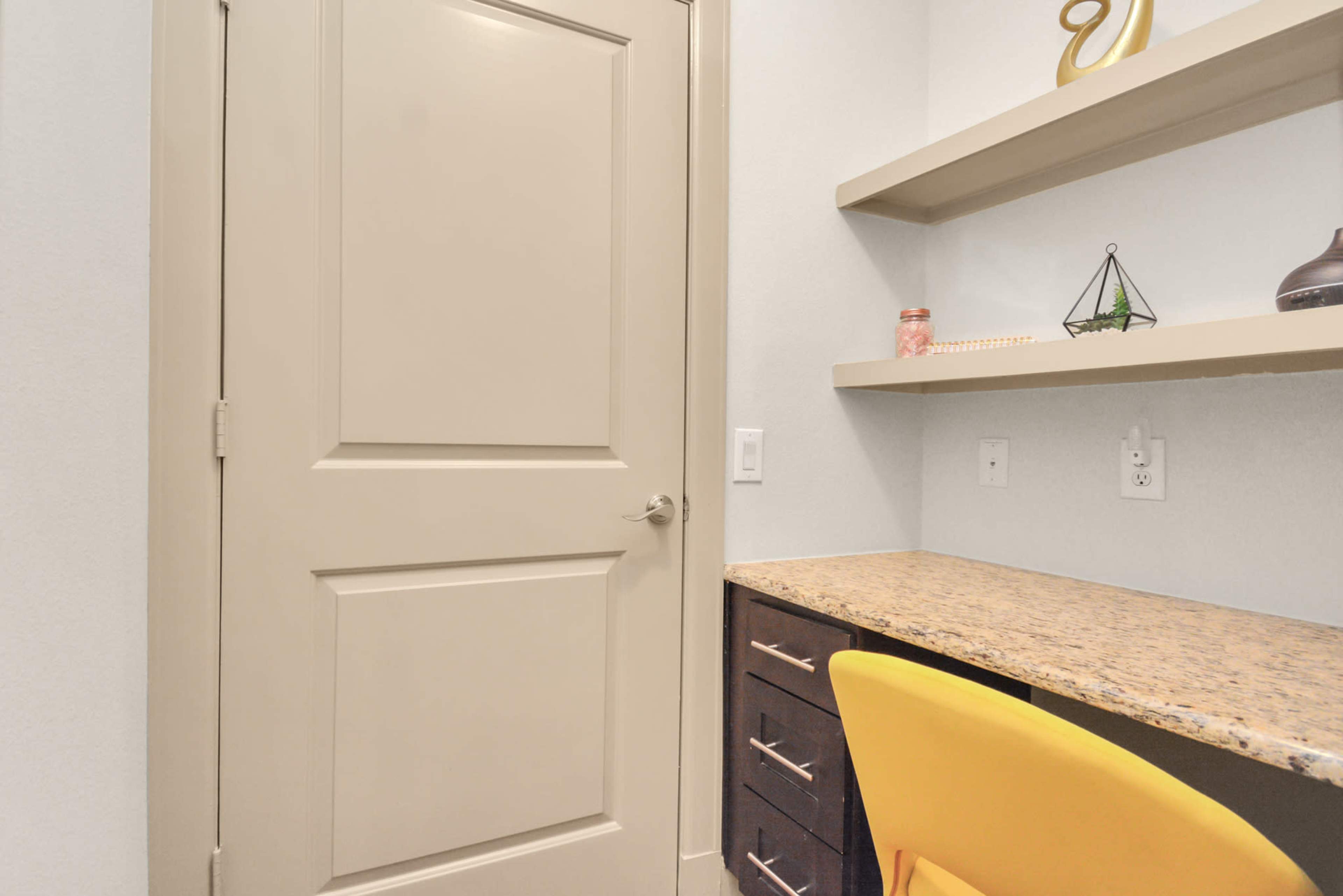 A small workspace with a granite-topped desk, a yellow chair, and a wall-mounted shelf, next to a closed beige door.