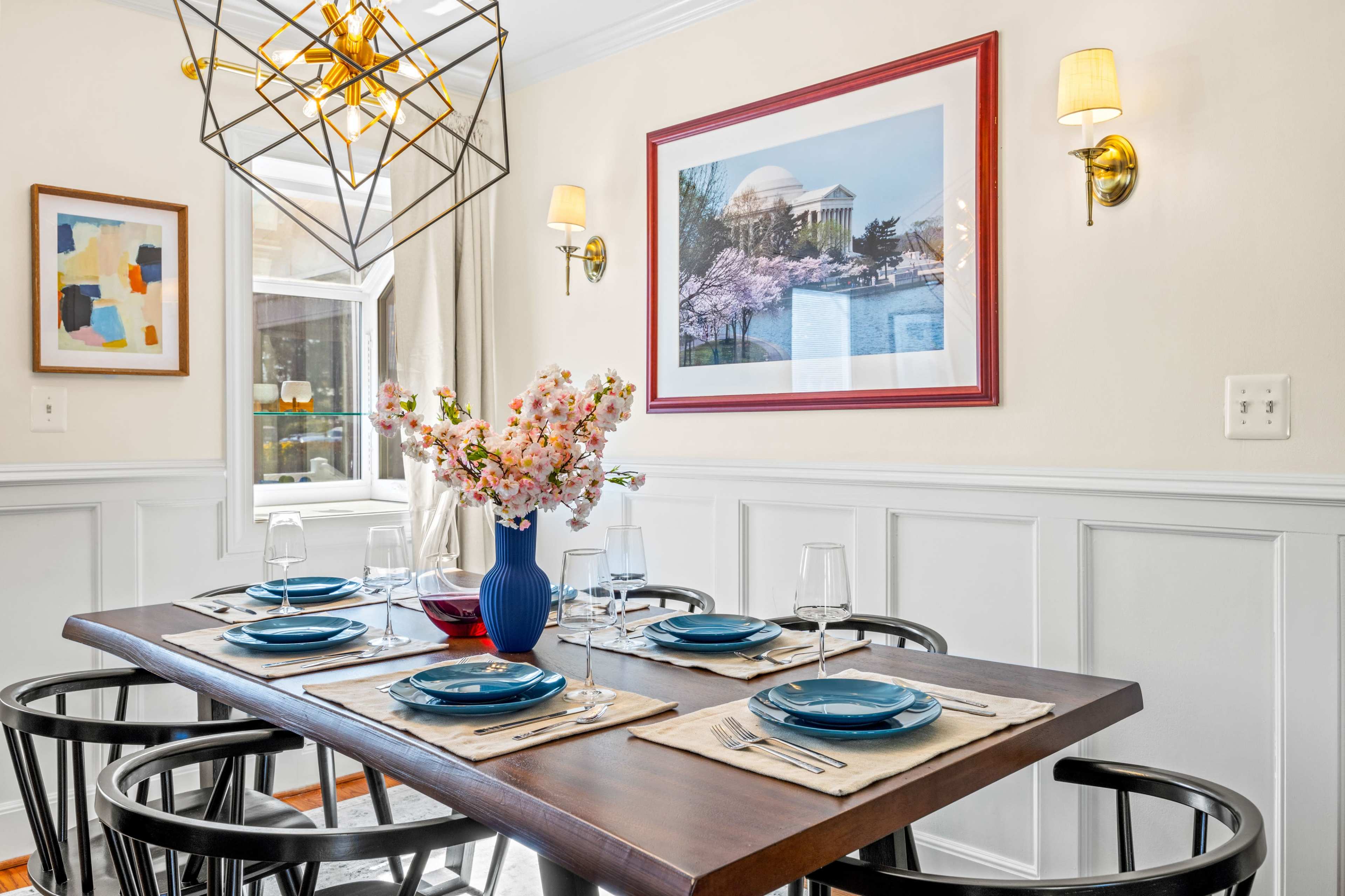 A dining table is set with blue plates and wine glasses, surrounded by black chairs, with a floral centerpiece and framed artwork on the wall.