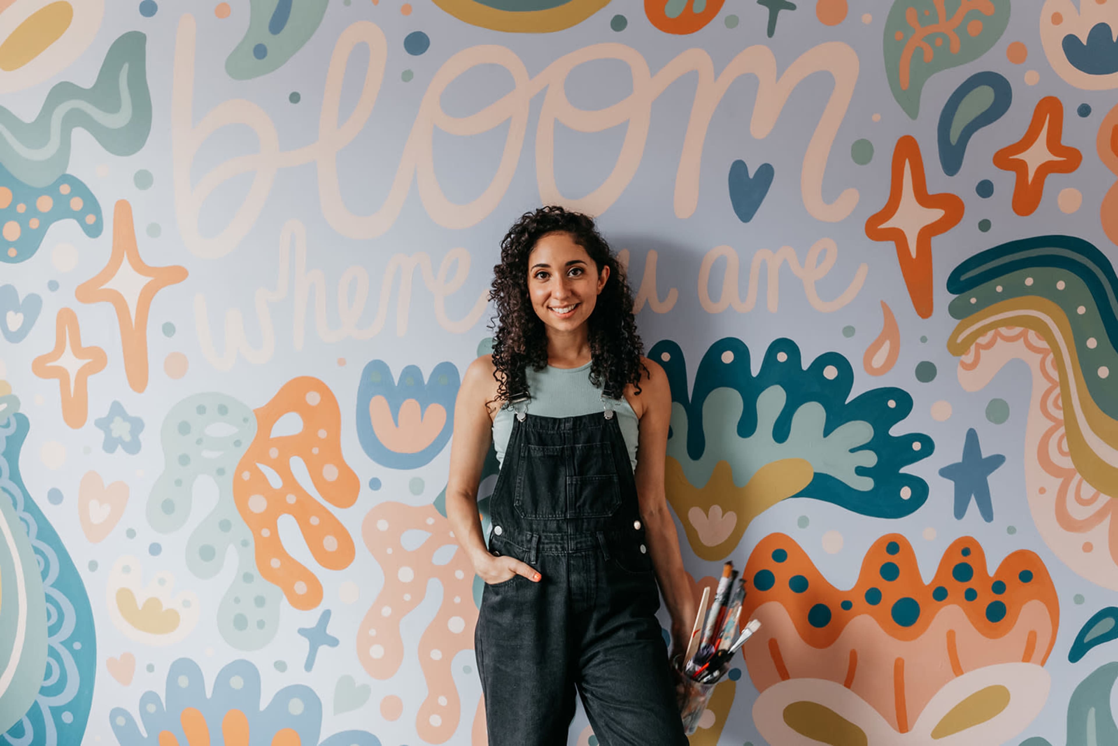A young woman with curly hair stands against a colorful mural that reads "bloom where you are," holding a bundle of paintbrushes.