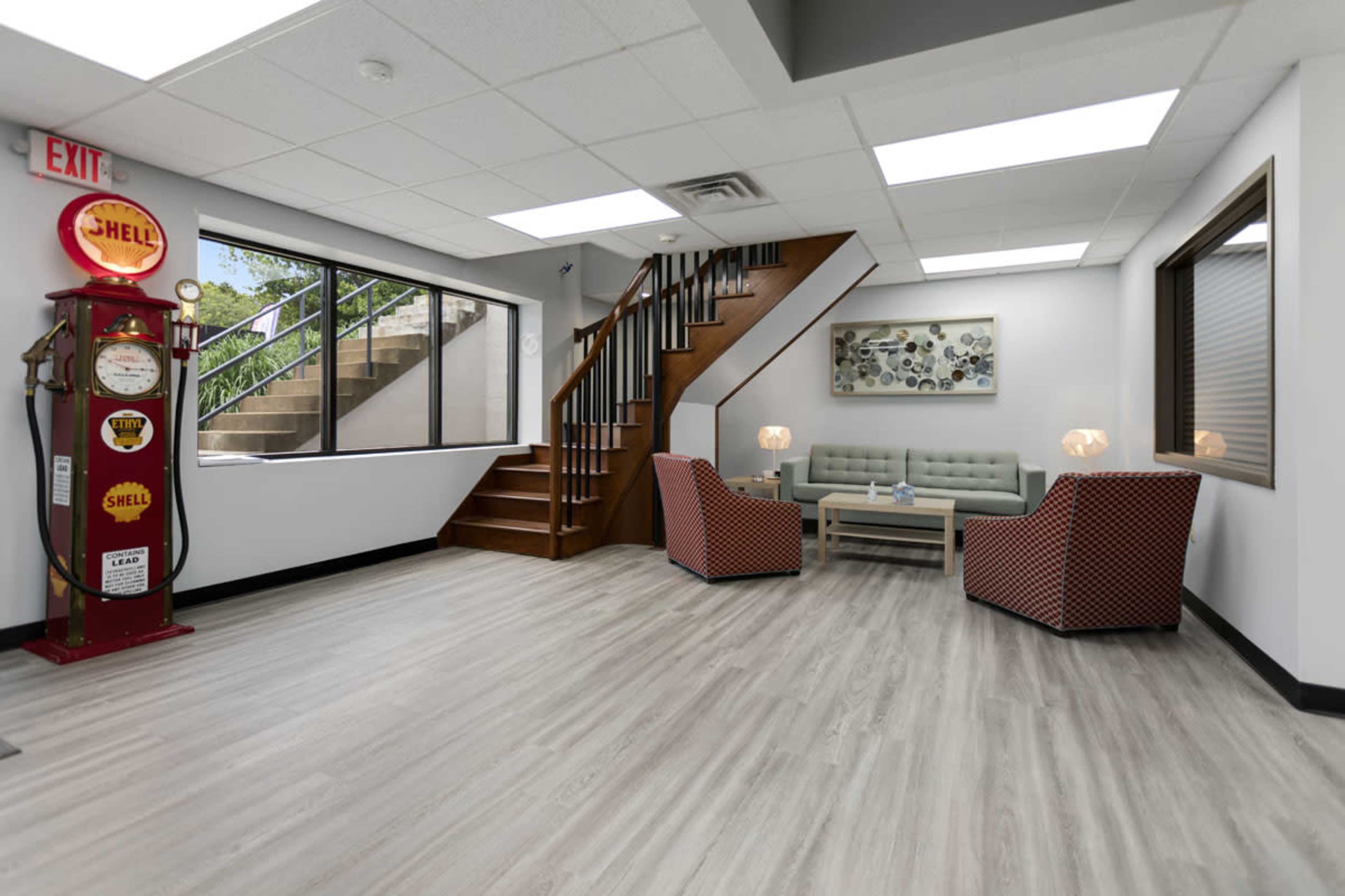 An interior space featuring a seating area with two patterned chairs and a light green couch, along with a staircase, a vintage Shell gas pump, and ample natural light coming from large windows.