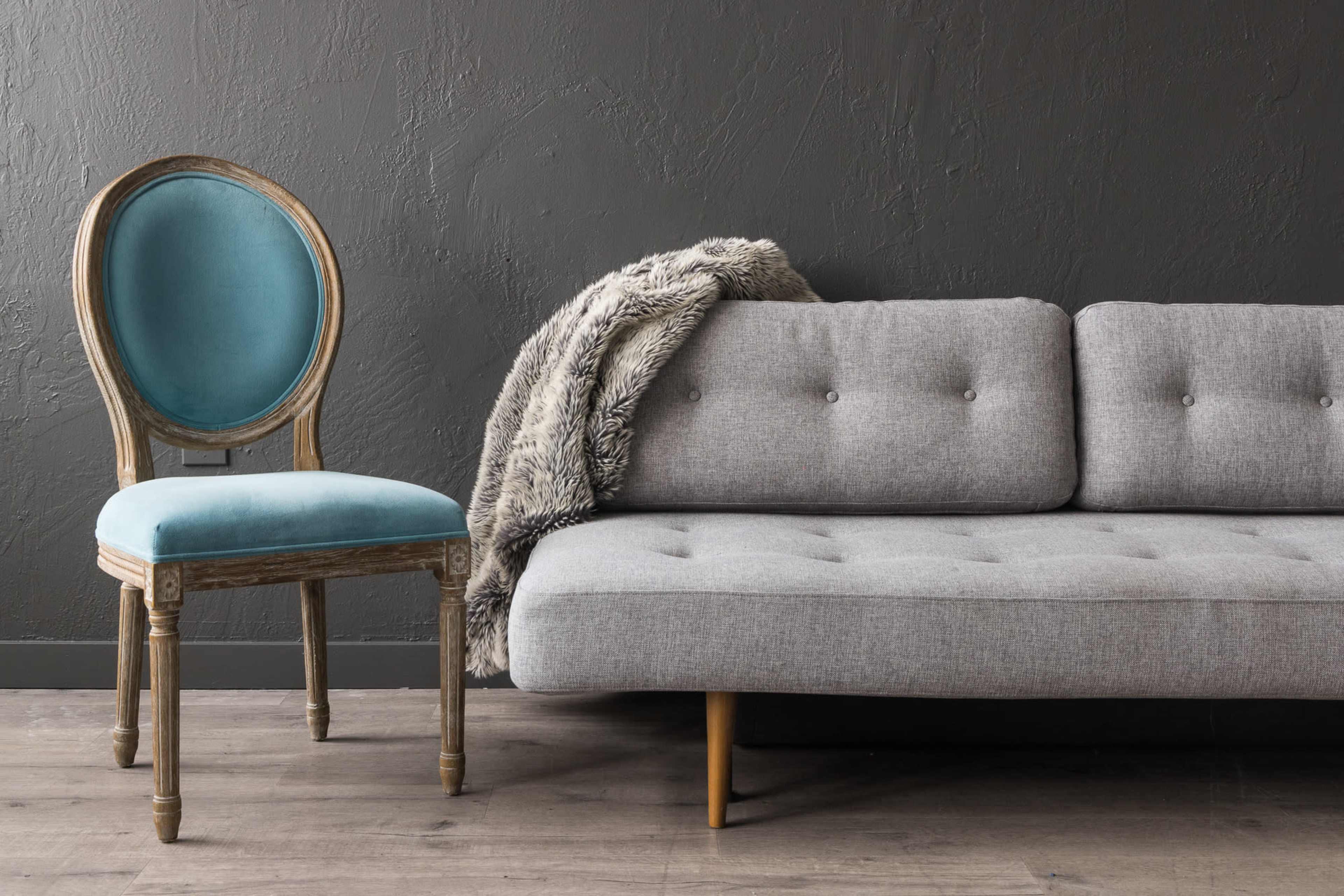 A gray sofa is positioned next to a vintage-style chair with a turquoise seat, against a dark textured wall.
