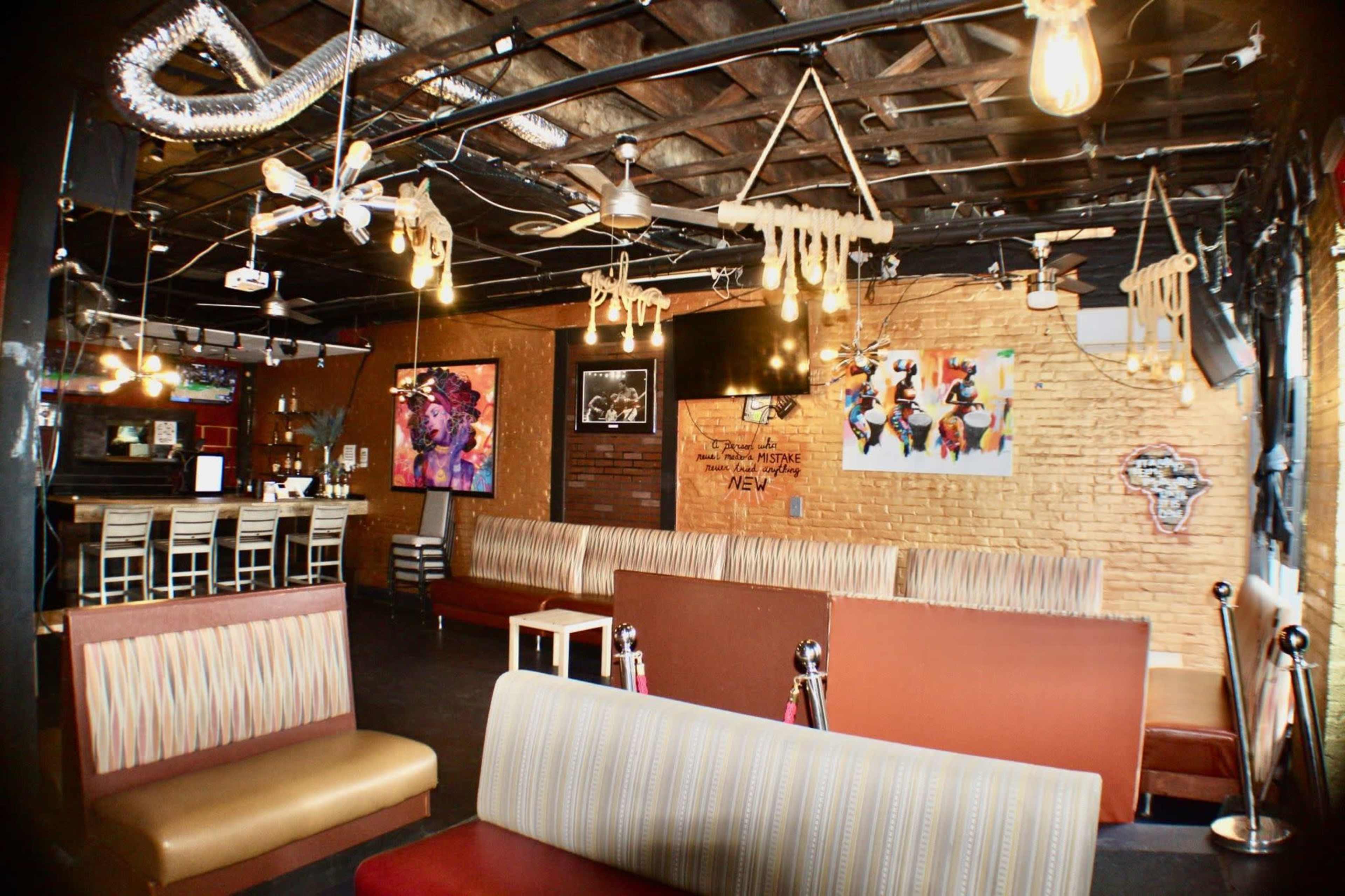 The image shows a spacious, dimly-lit interior of a bar or lounge featuring various seating arrangements, exposed brick walls, and decorative art on the walls.
