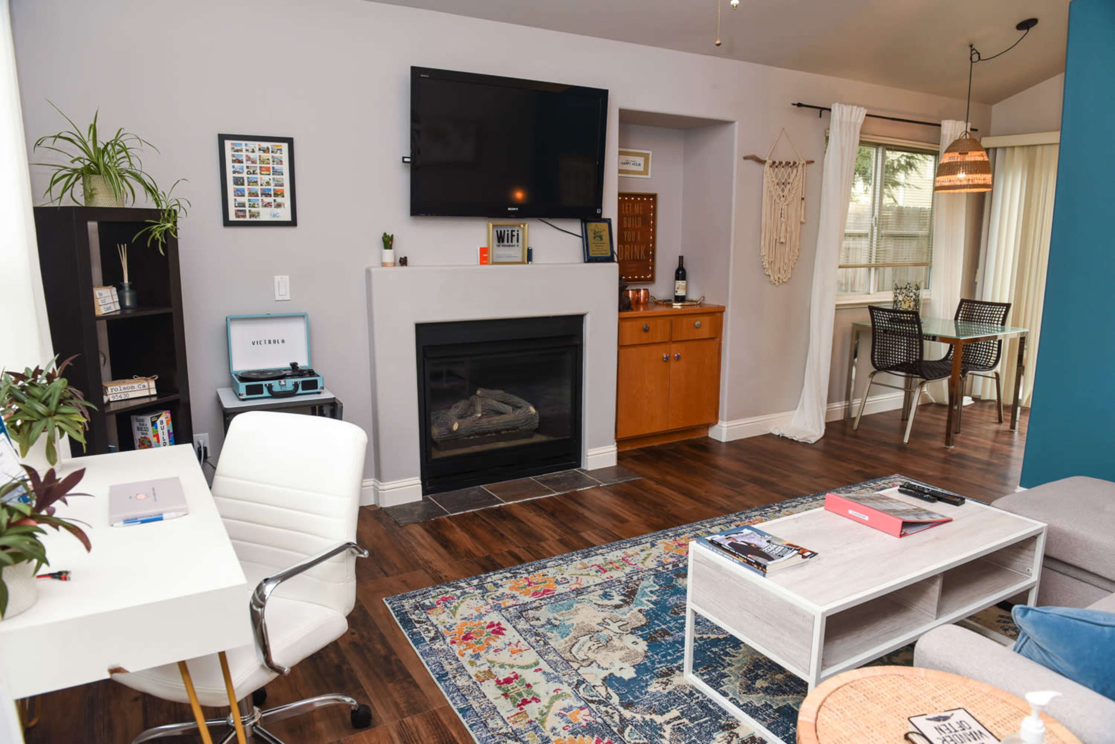 The image shows a cozy living room with a fireplace, television mounted on the wall, a small dining area, and a desk workspace.
