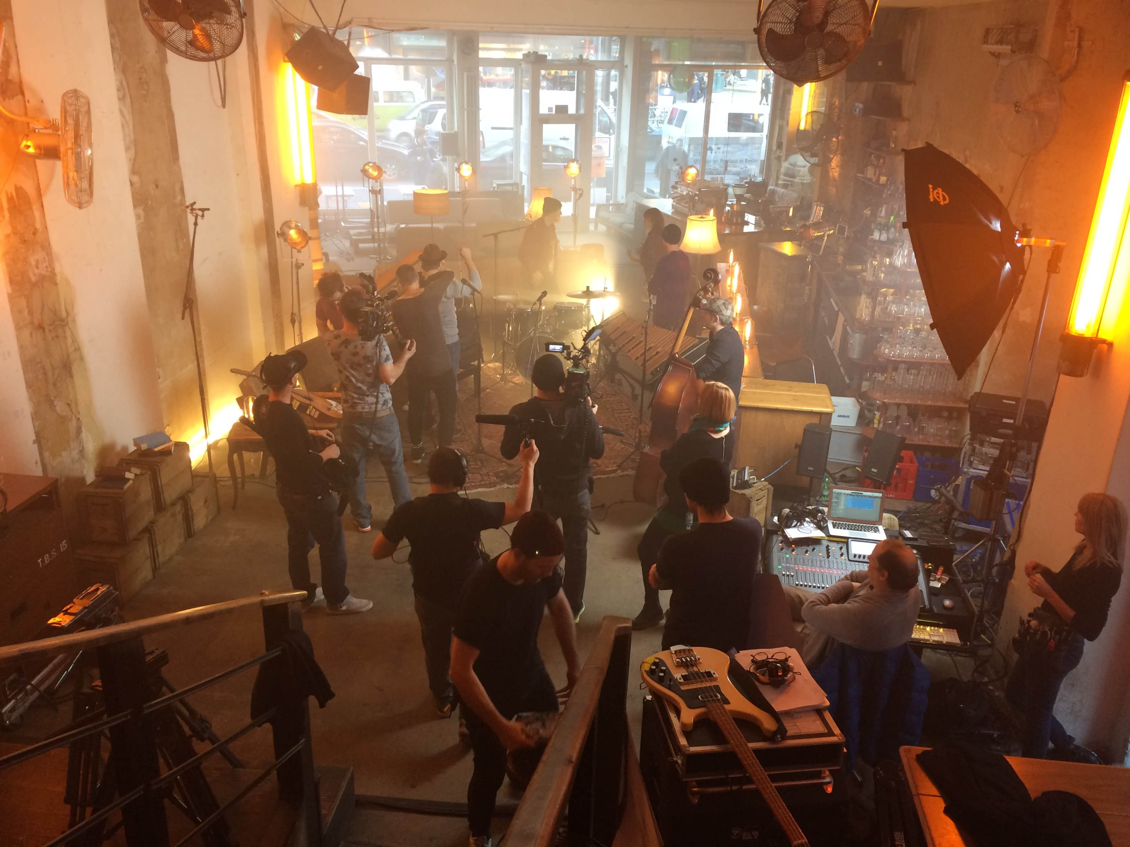 A band performs in a lively venue with microphones and cameras set up around them, while crew members manage sound and lighting.