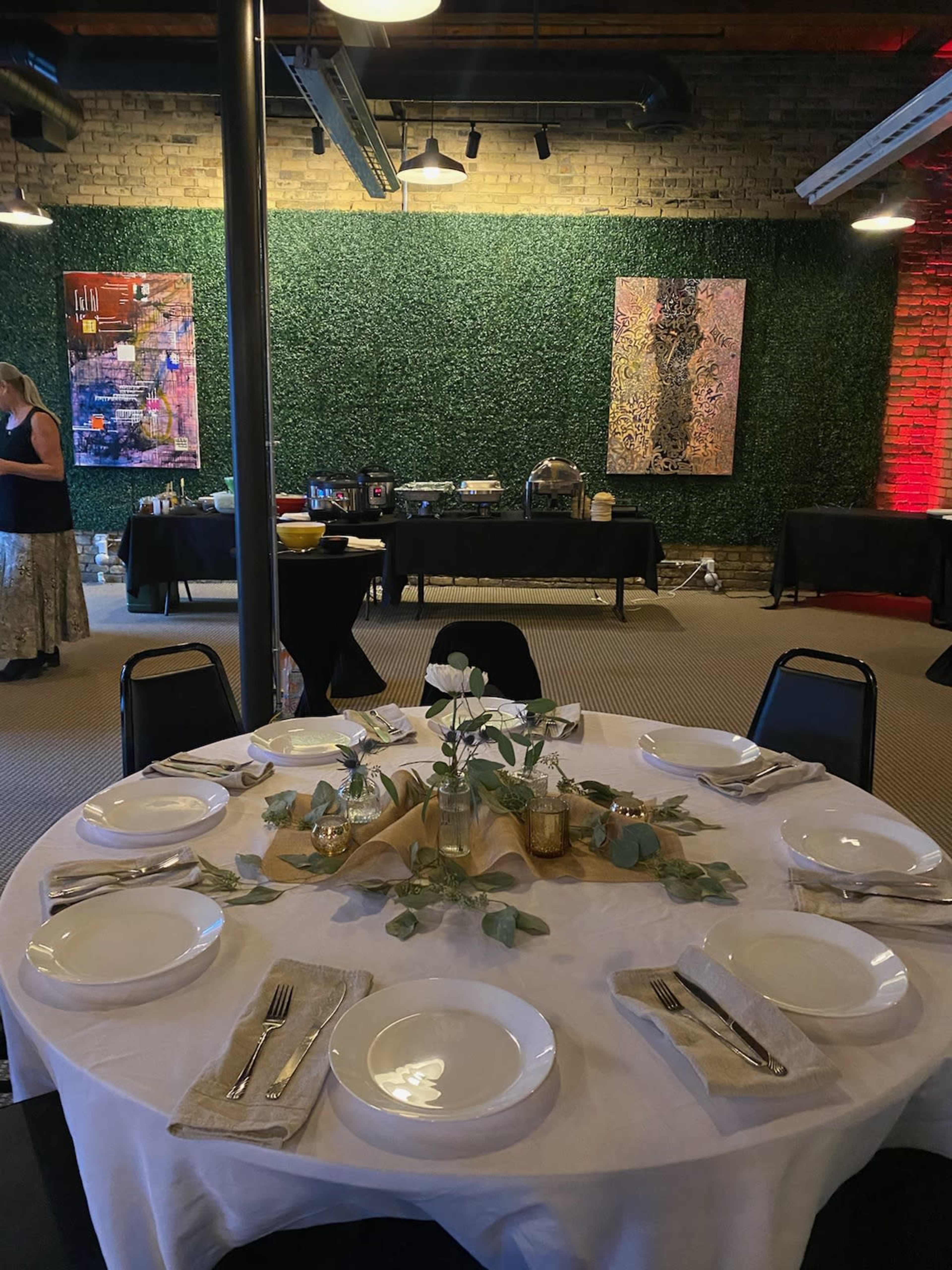 A round table is set with plates, utensils, and a floral centerpiece in a decorated venue featuring green wall art and food stations in the background.