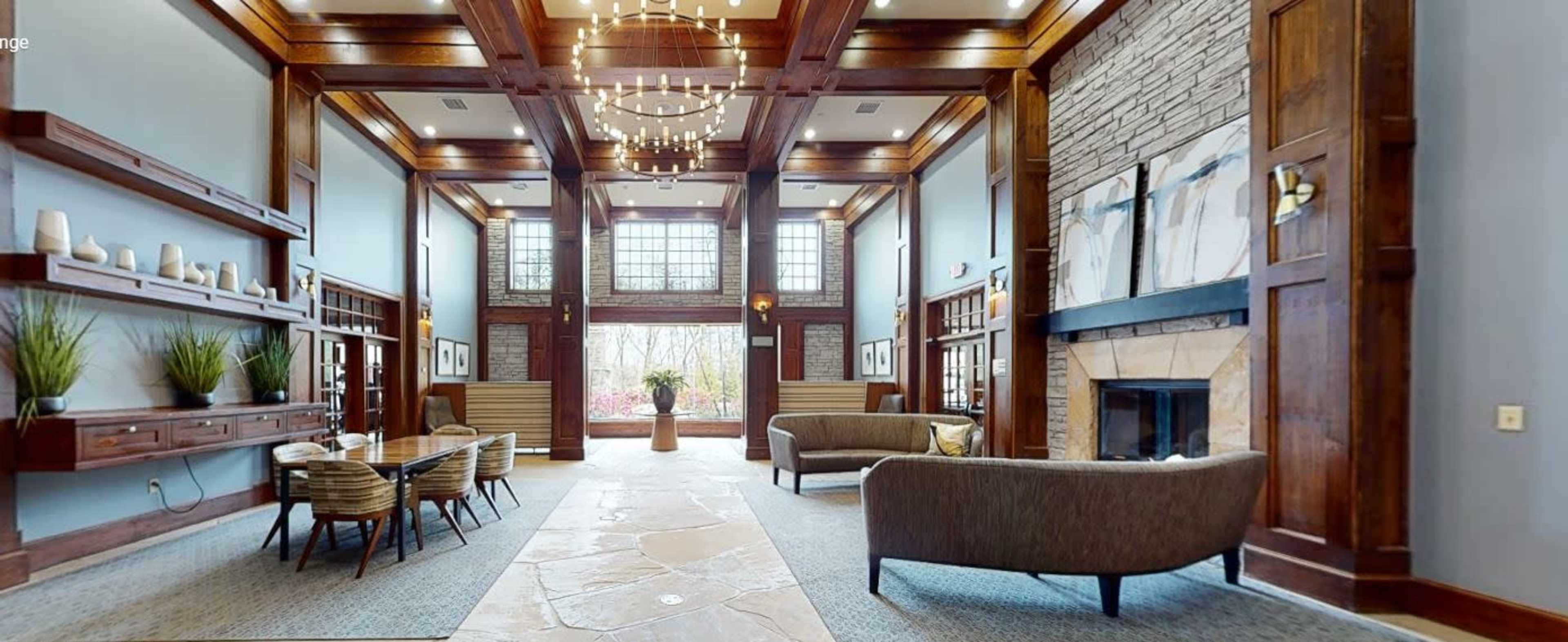 The image shows a spacious lobby interior featuring wooden pillars, a stone fireplace, modern seating arrangements, and large windows allowing natural light to fill the space.