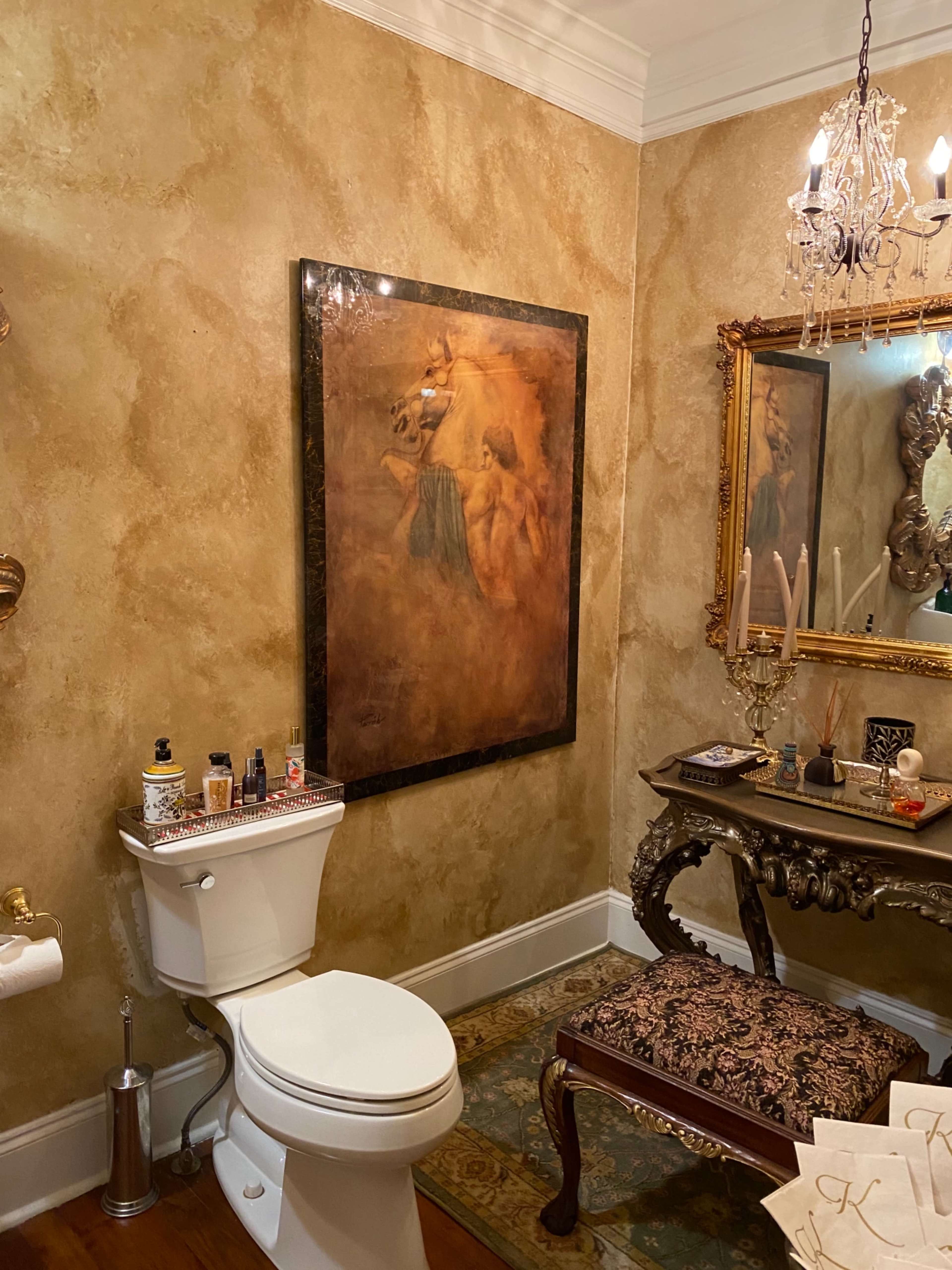 A bathroom with a toilet, a decorative table, an ornate mirror, and a large painting on the wall.