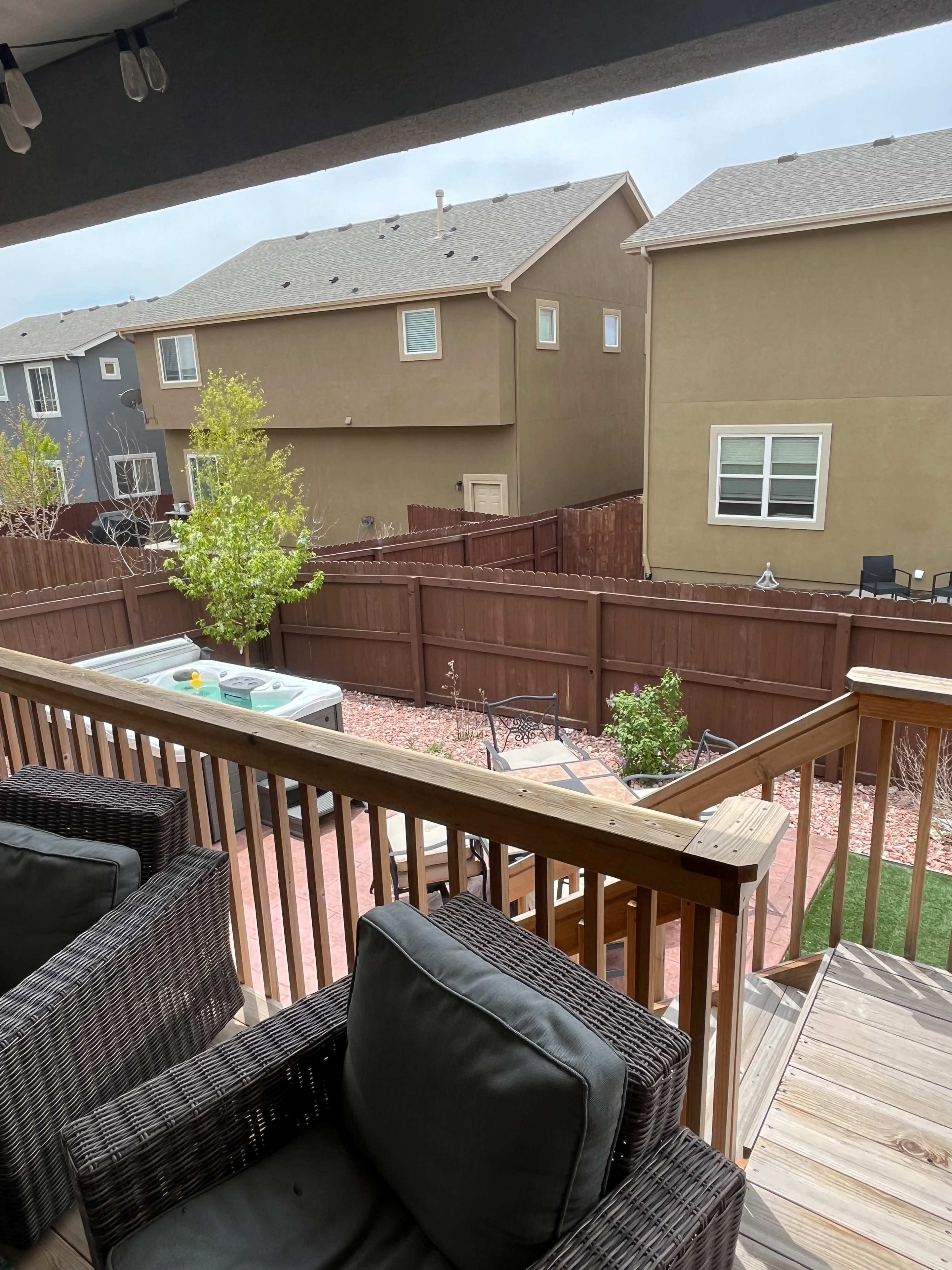 The image shows a backyard view from a deck, featuring a hot tub, outdoor furniture, and a fenced area with neighboring houses in the background.