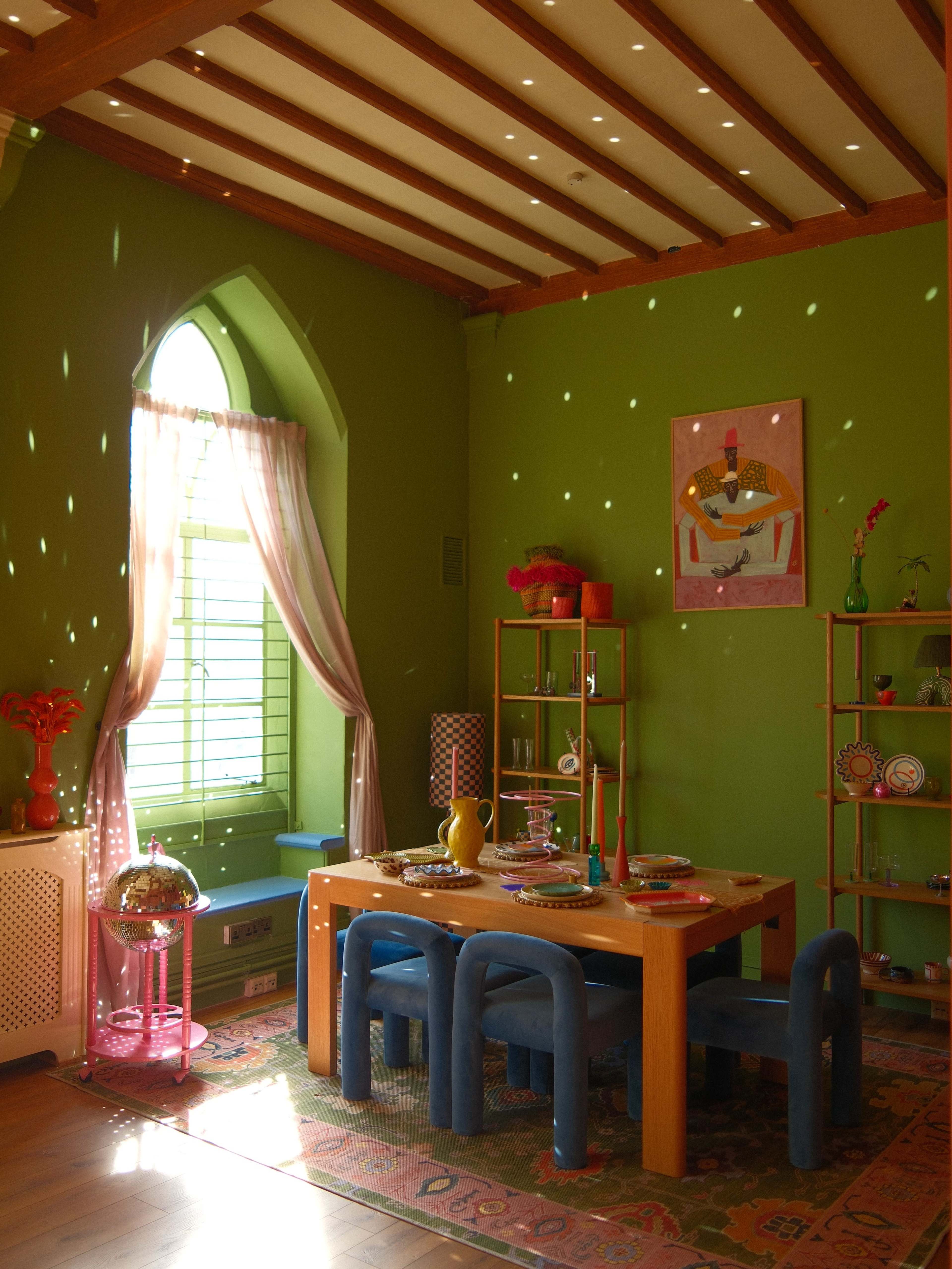 The image shows a brightly lit dining area featuring a green wall, a wooden table surrounded by blue chairs, and decorative items on shelves and the table.