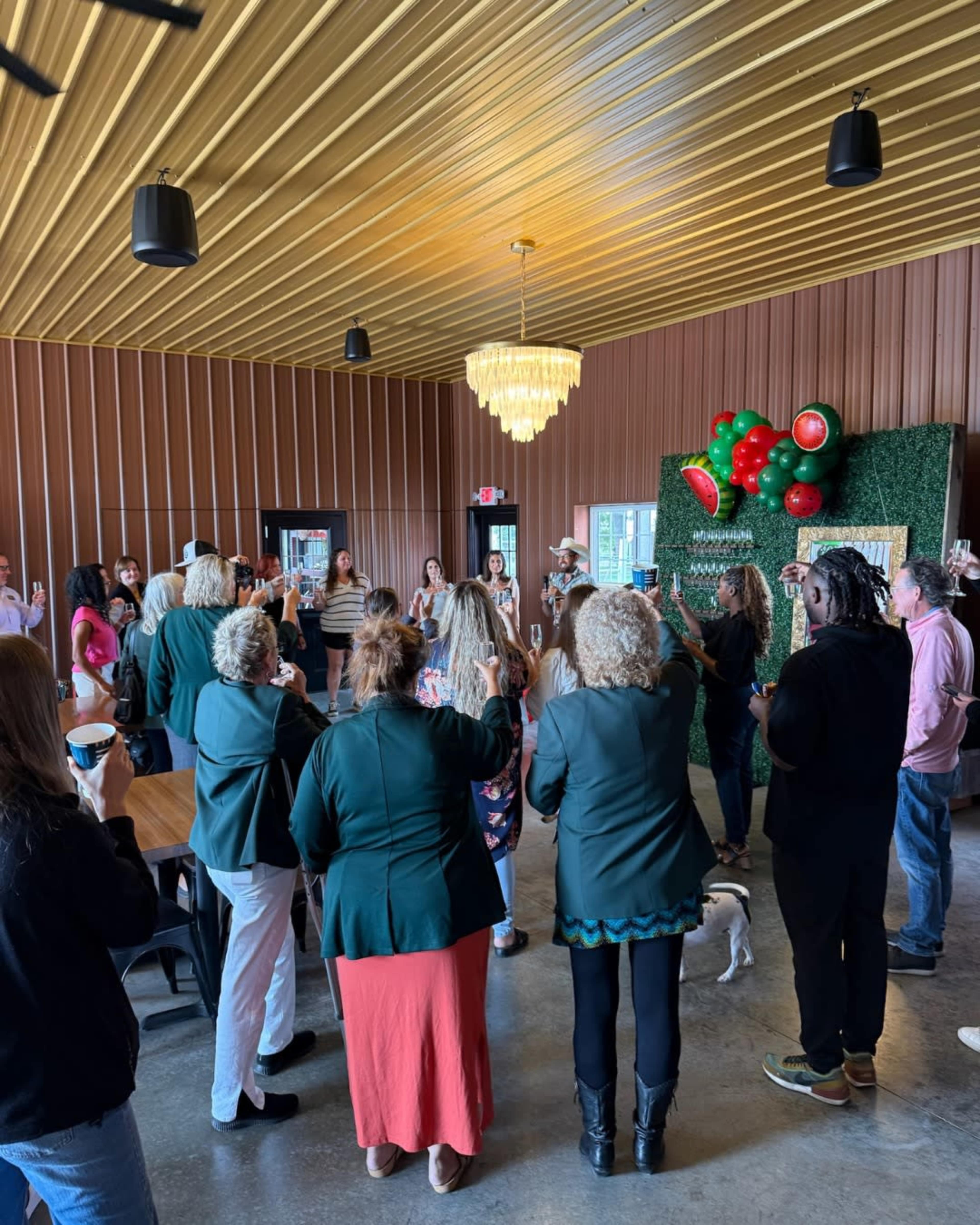 A group of people is gathered indoors, participating in an event with a decorative backdrop featuring greenery and red accents.