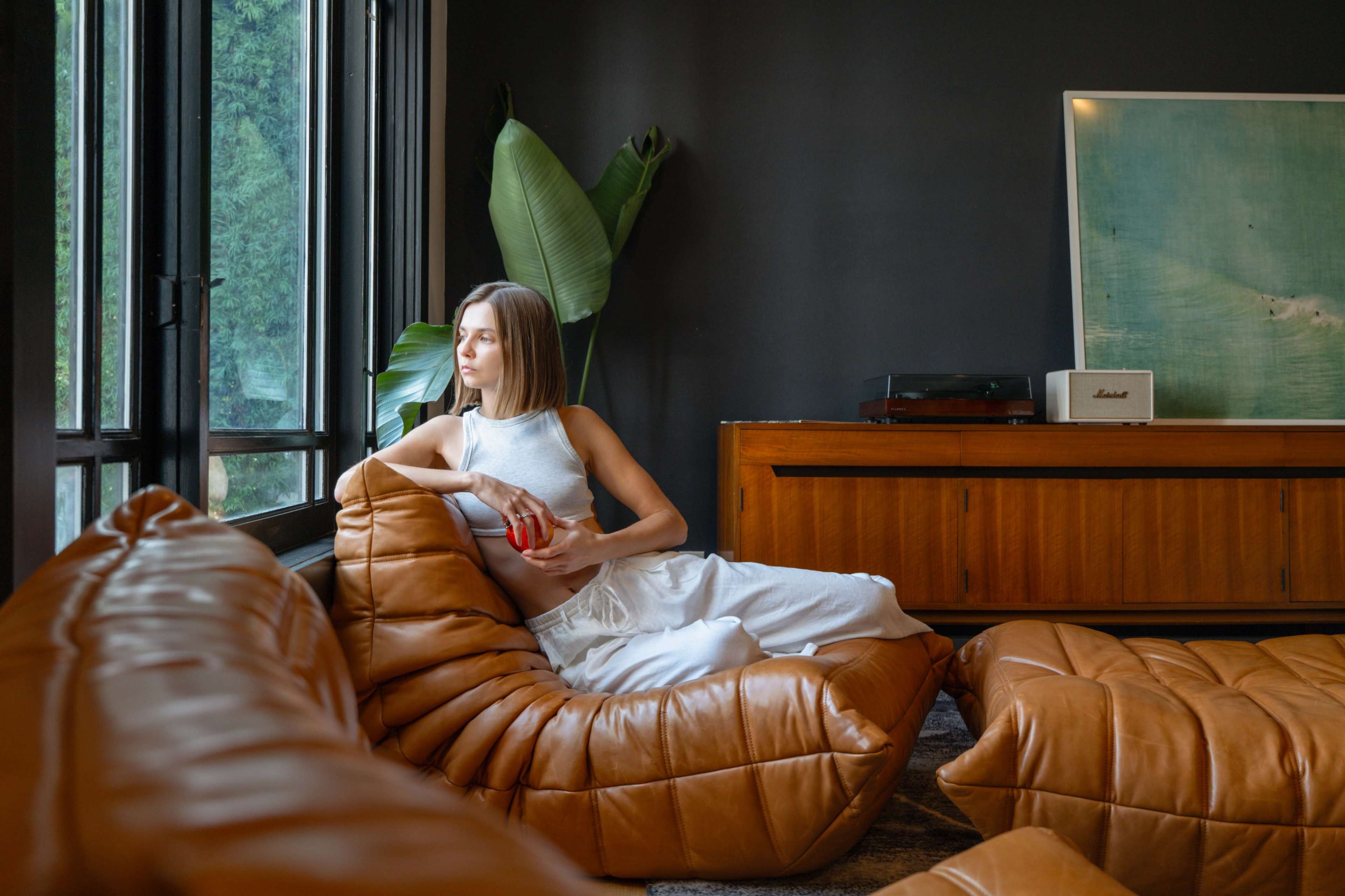 A woman sits on a brown leather couch in a modern living room, gazing out a large window beside a potted plant.