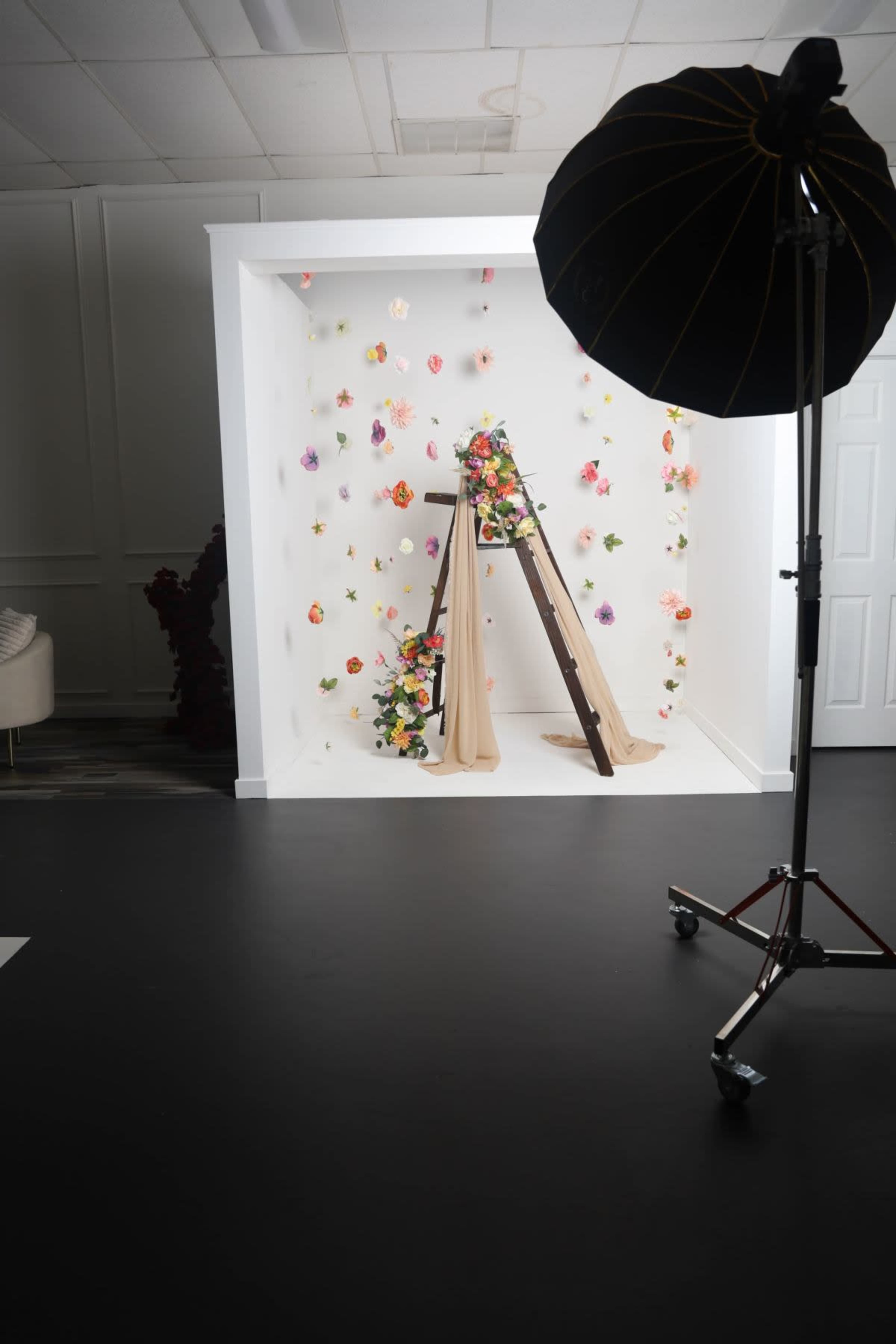 A photo studio setup featuring a white backdrop adorned with colorful floral decorations, a wooden ladder draped with fabric, and a large light stand positioned nearby.