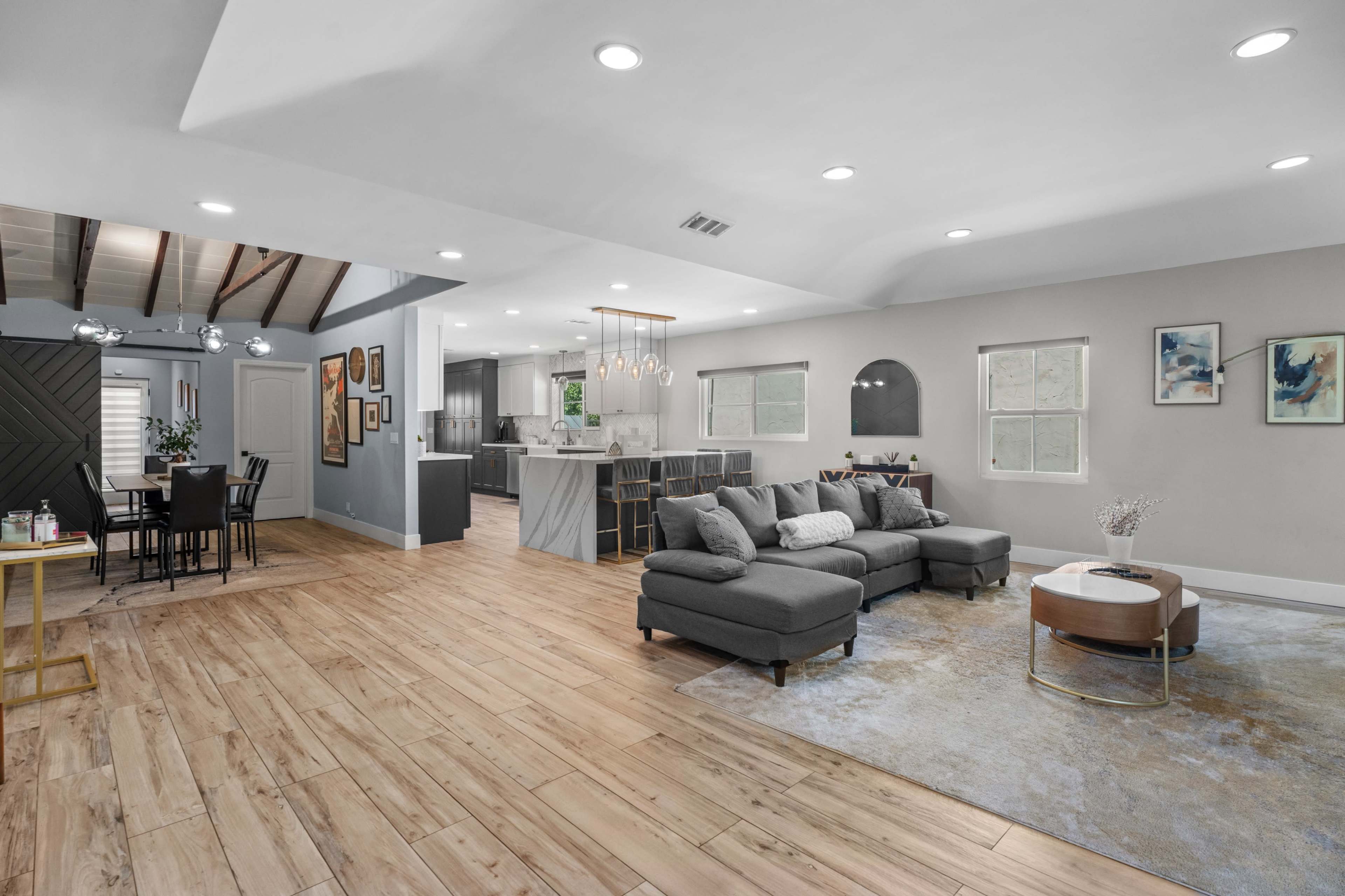 The image shows a modern, open-concept living space featuring a gray sectional sofa, a round coffee table, and a dining area with a table and chairs, all set against light wood flooring and neutral-colored walls.