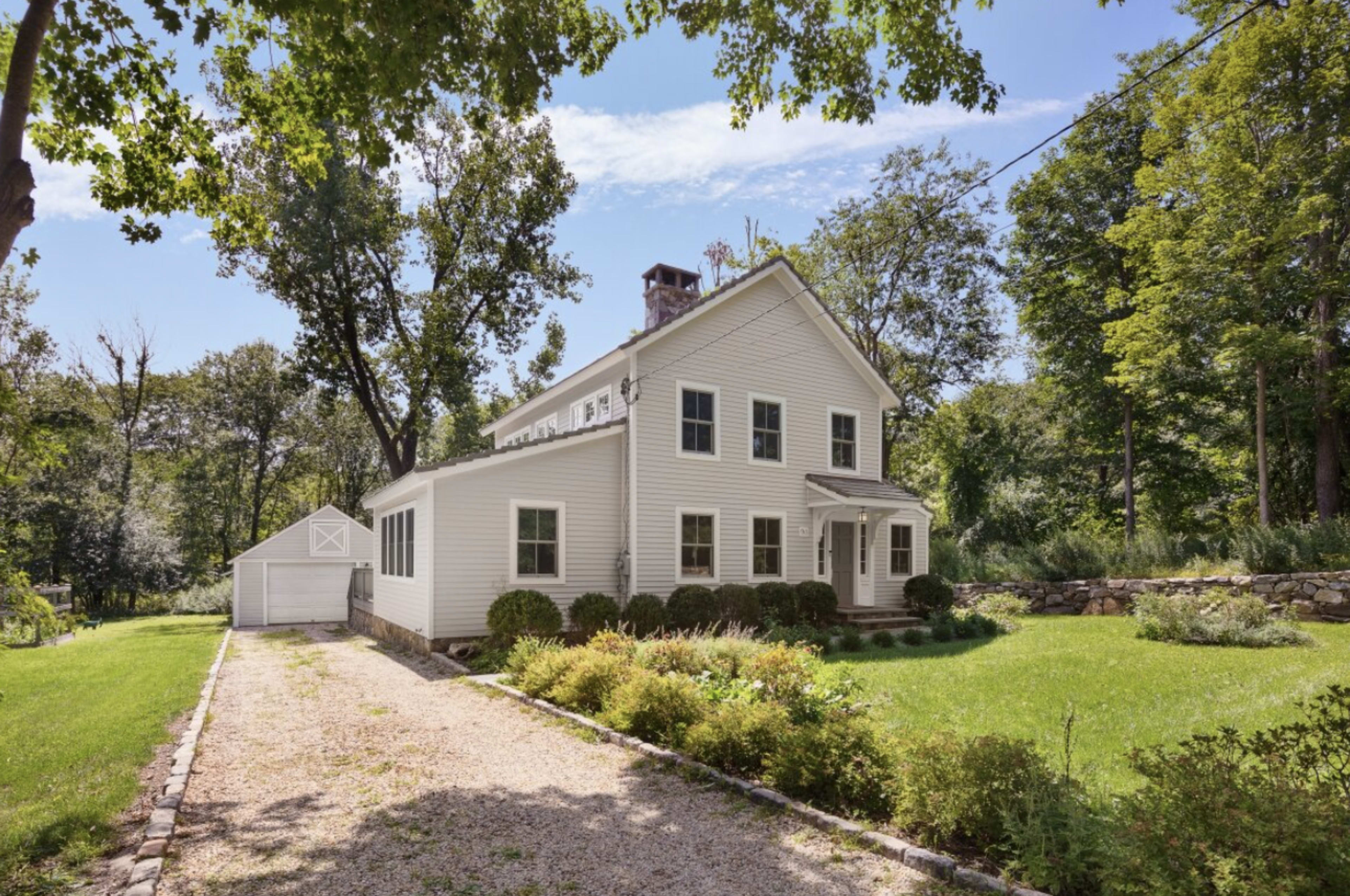 A two-story white house with a gravel driveway and a garage sits surrounded by trees and greenery.