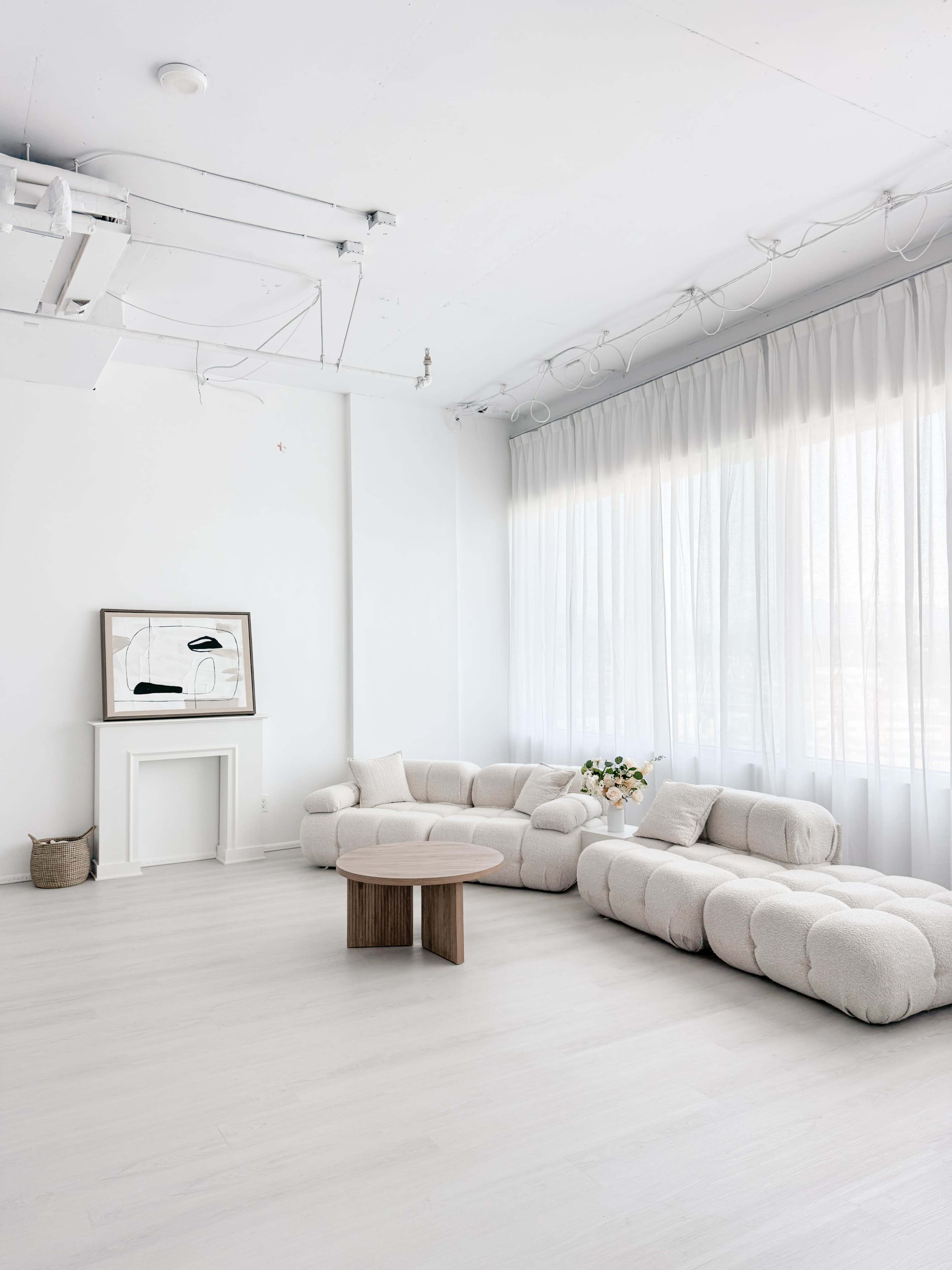 The image shows a modern, minimalist living room featuring a curved beige sofa, a round wooden coffee table, and large windows with sheer white curtains.