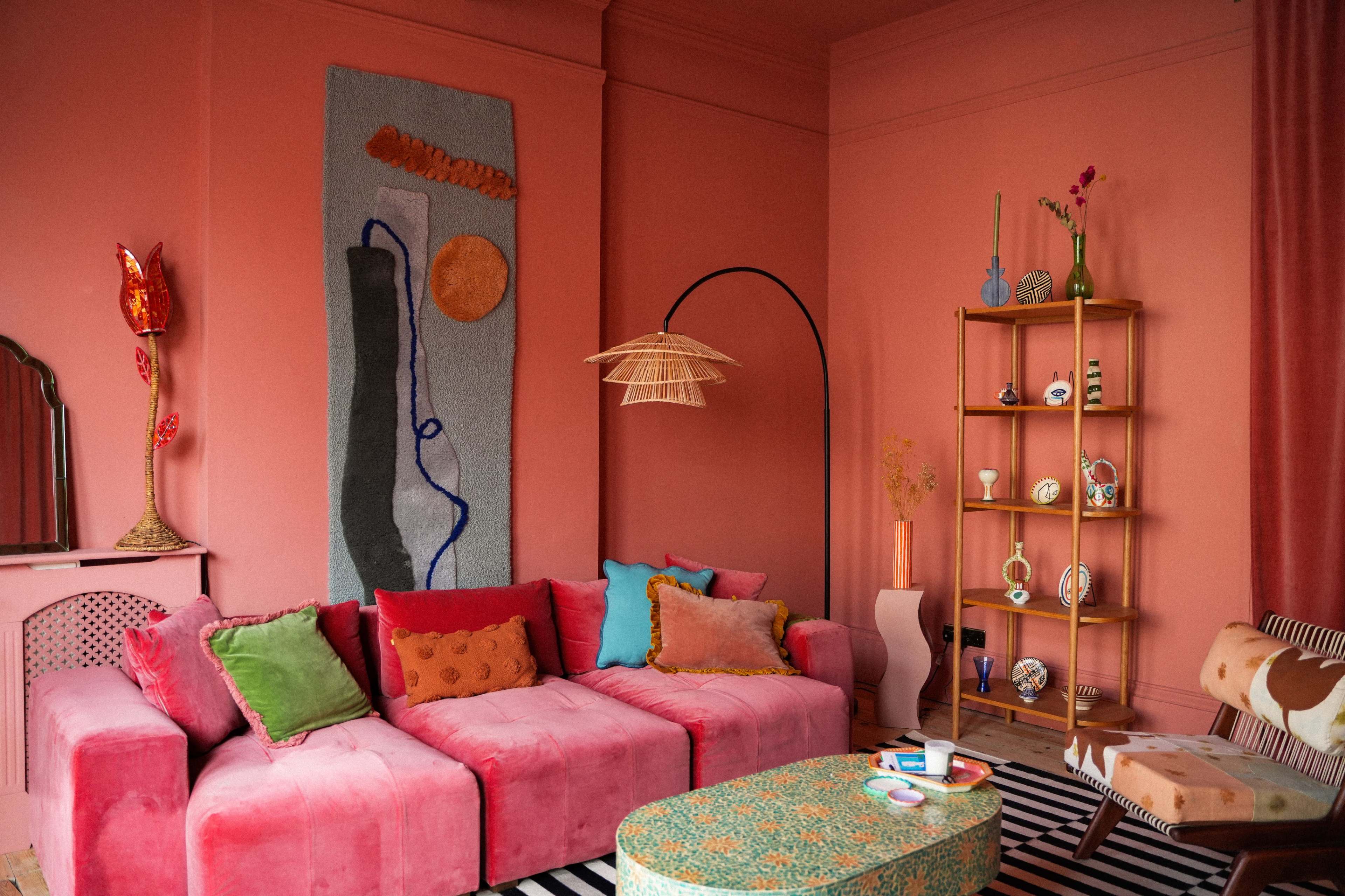The living room features a pink sectional sofa adorned with colorful cushions, a patterned coffee table, and a shelf displaying various decorative items against a matching pink wall.