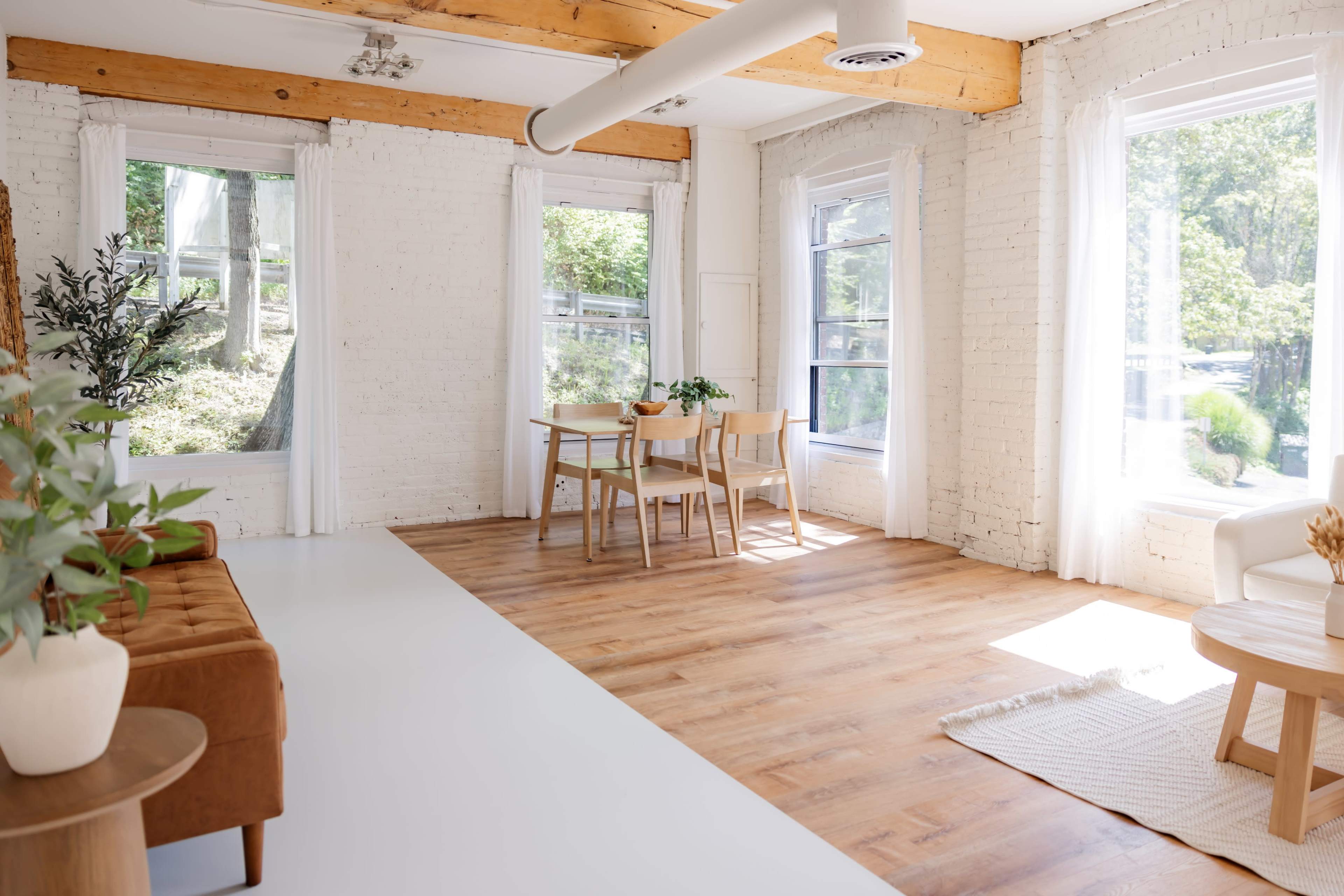 The image shows a bright and airy room with large windows, a wooden table and chairs, a sofa, and plants, featuring light-colored walls and wooden beams.