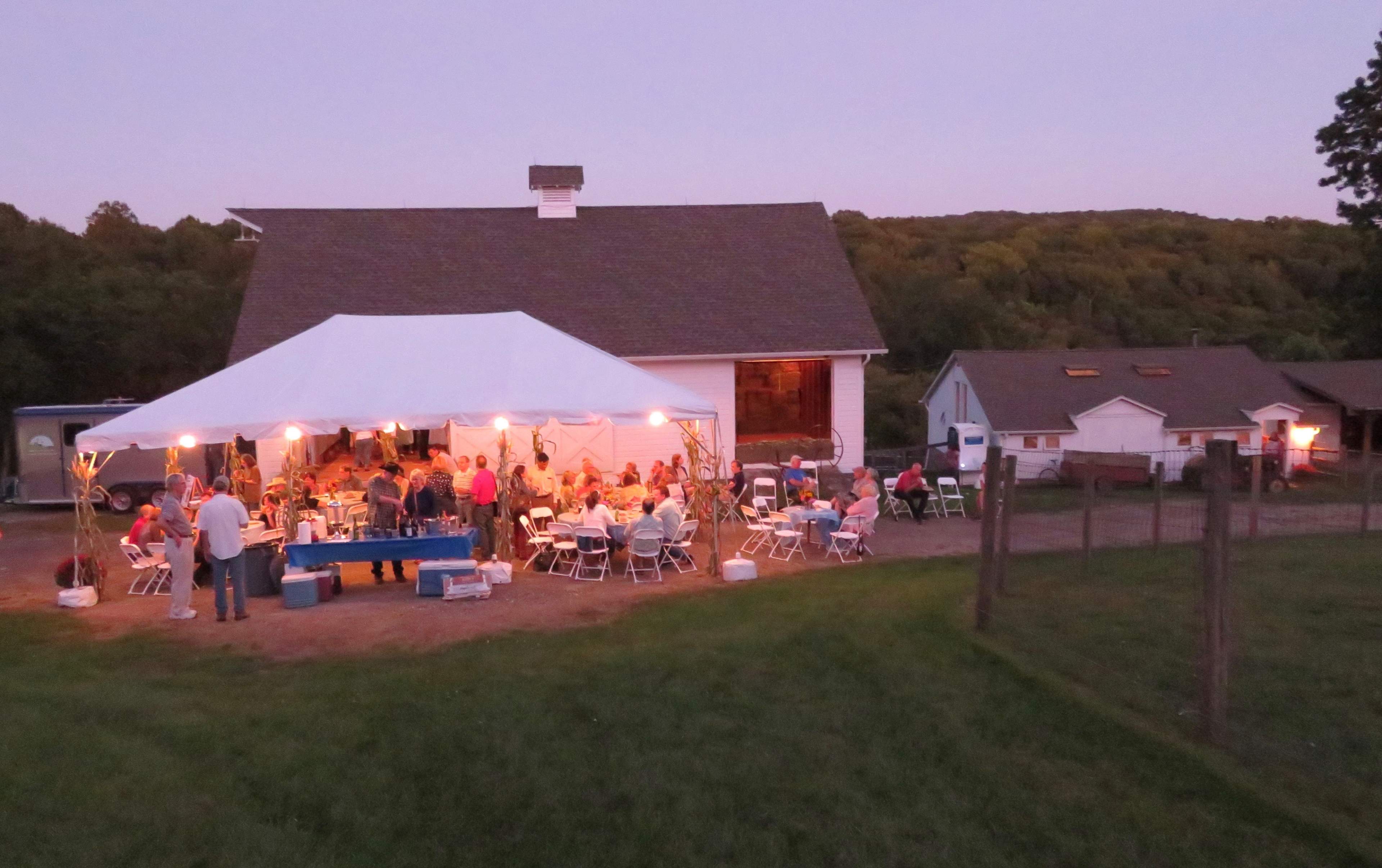 A large white tent with chairs and tables is set up outside a barn, with people gathered for an evening event as dusk falls.