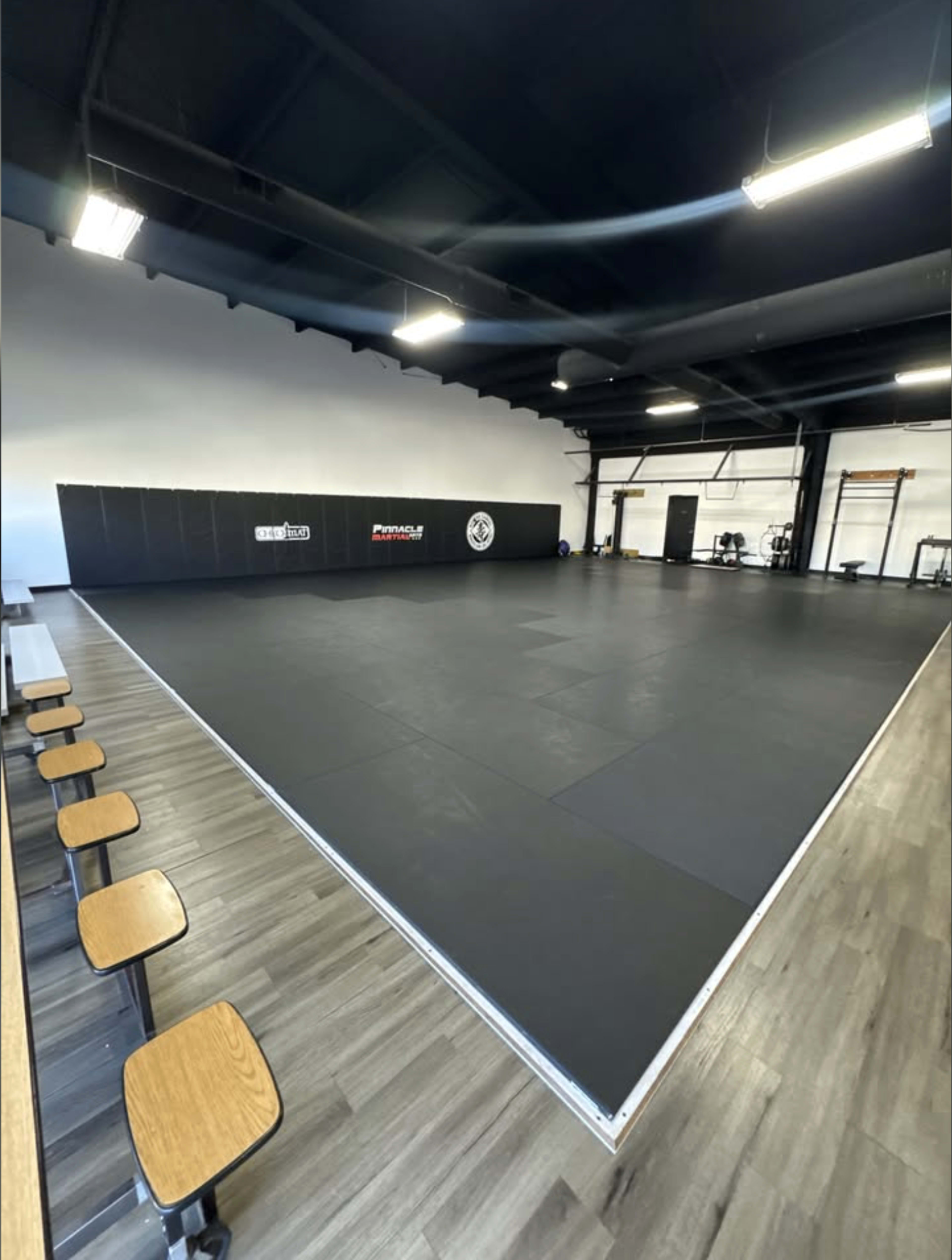 The image shows a spacious martial arts training area with black padded flooring and a seating section along one side.