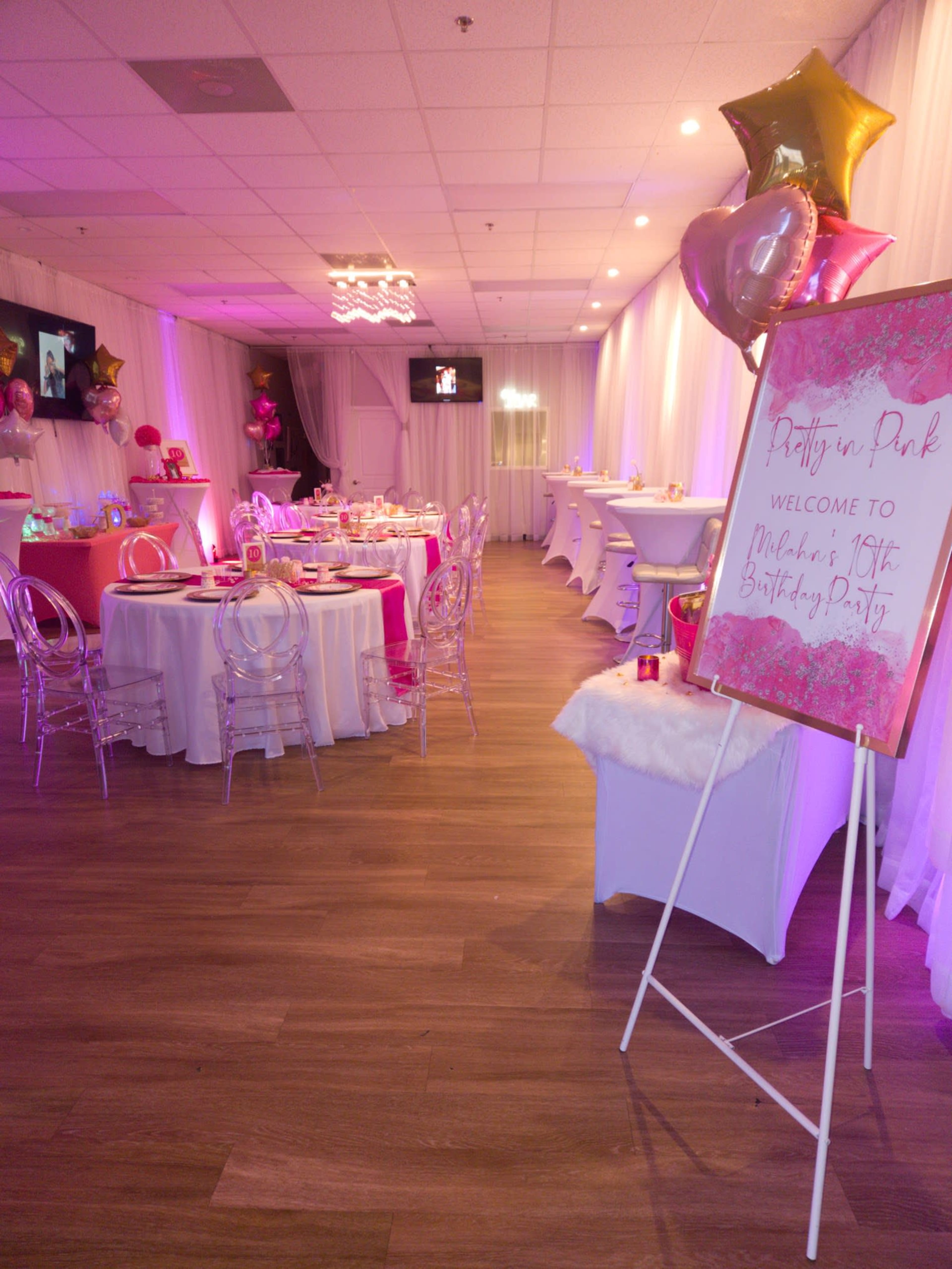 A brightly decorated indoor party space with tables set for a birthday celebration, featuring pink accents, balloons, and a welcoming sign.
