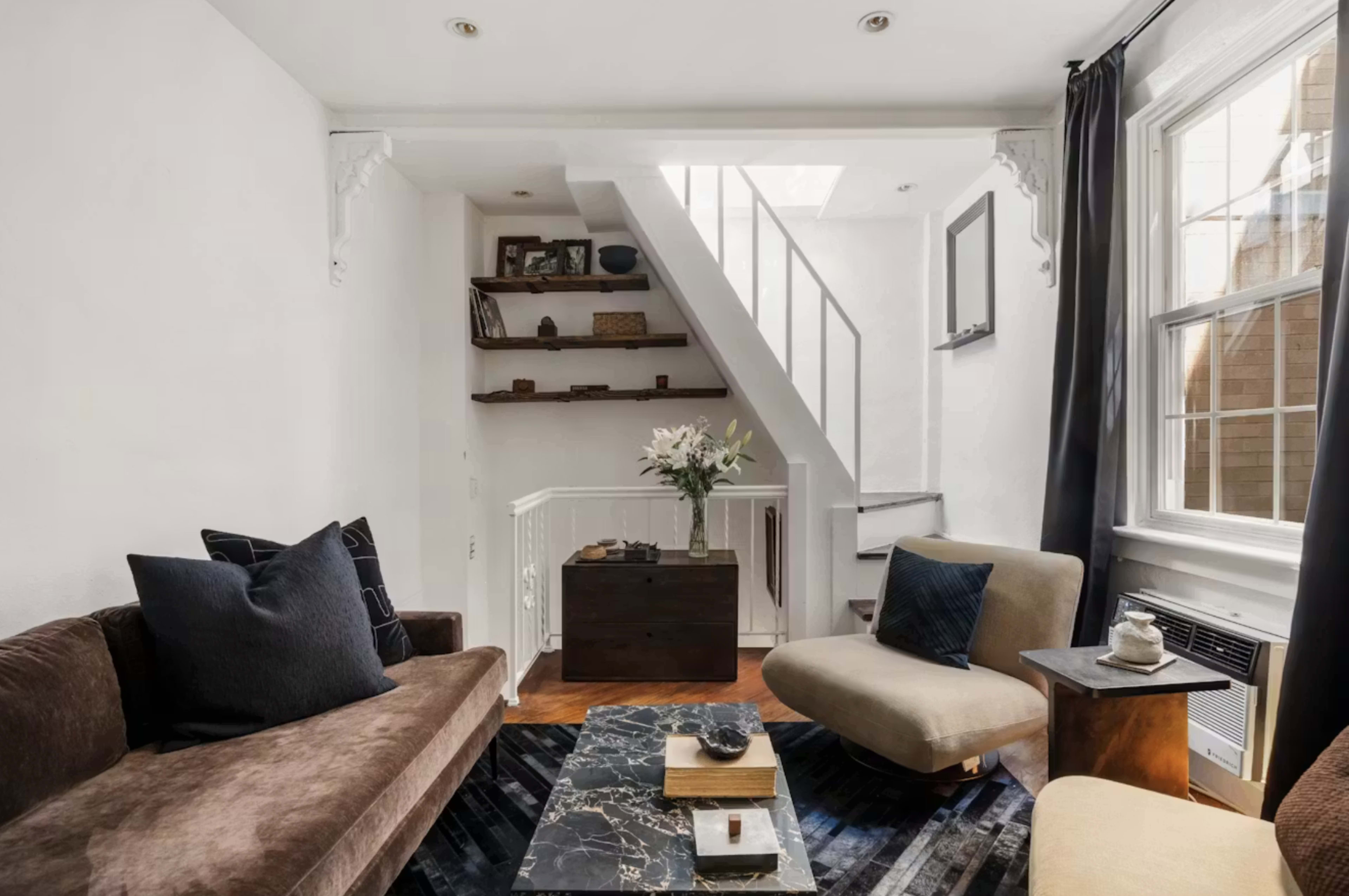 The image shows a modern living room with a brown sofa, an armchair, a decorative coffee table, and a staircase leading to an upper level.