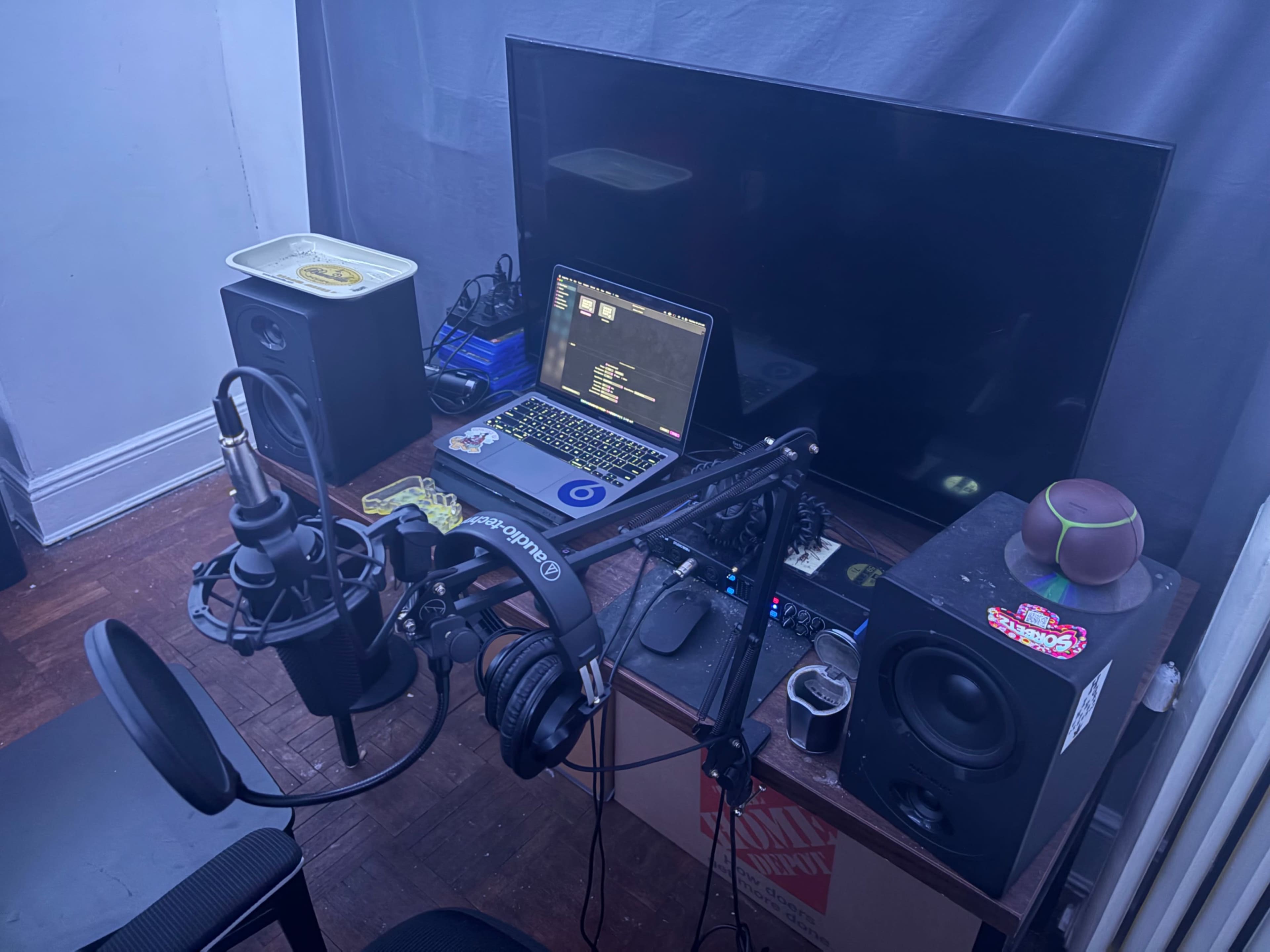 The image shows a recording setup featuring a microphone on an adjustable stand, a laptop on a desk, and various audio equipment, including a speaker and a coffee mug.