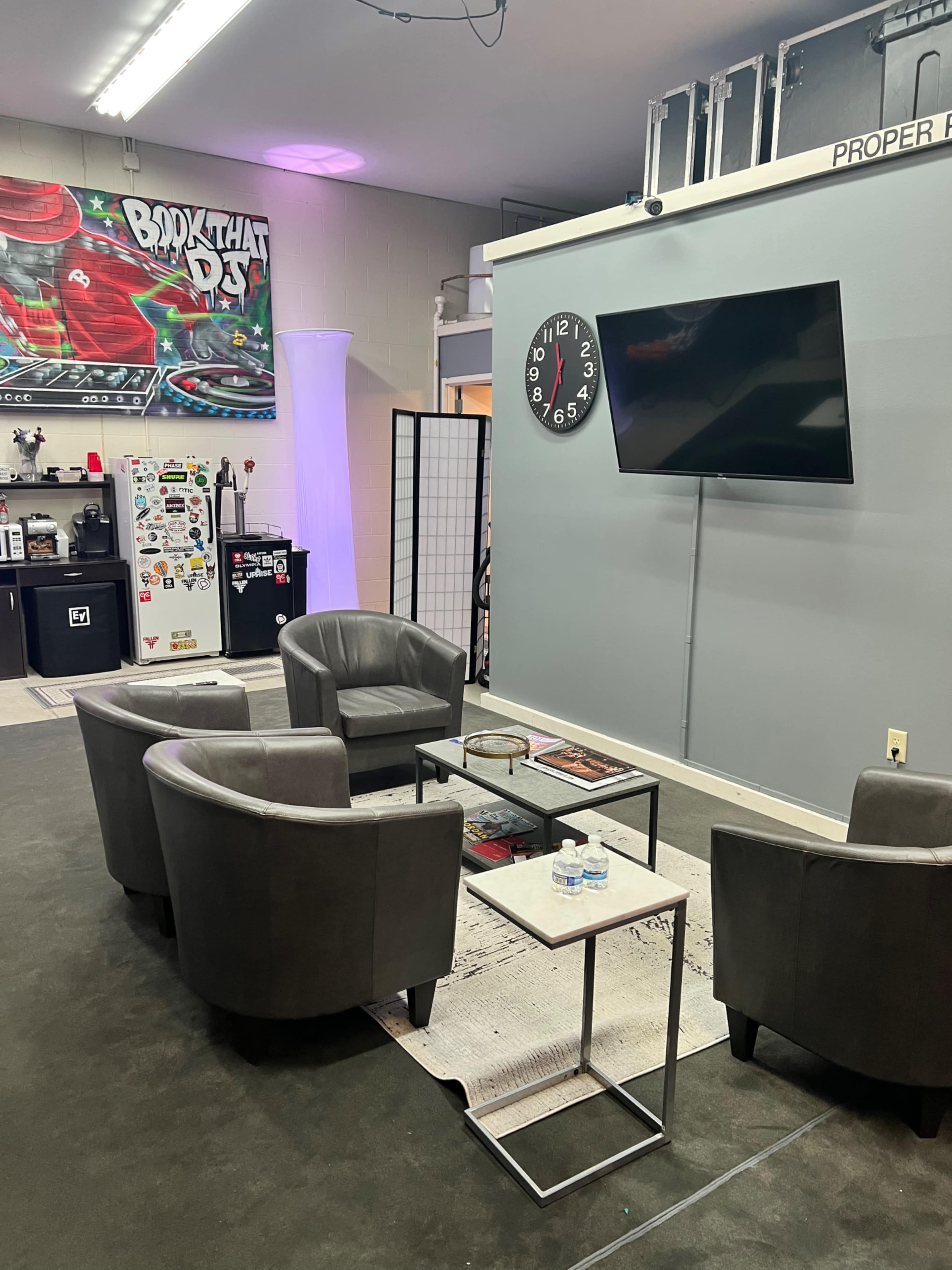 A seating area with four gray chairs and a small table is set against a wall featuring a large clock and a TV in a mixed-use space decorated with a colorful mural.
