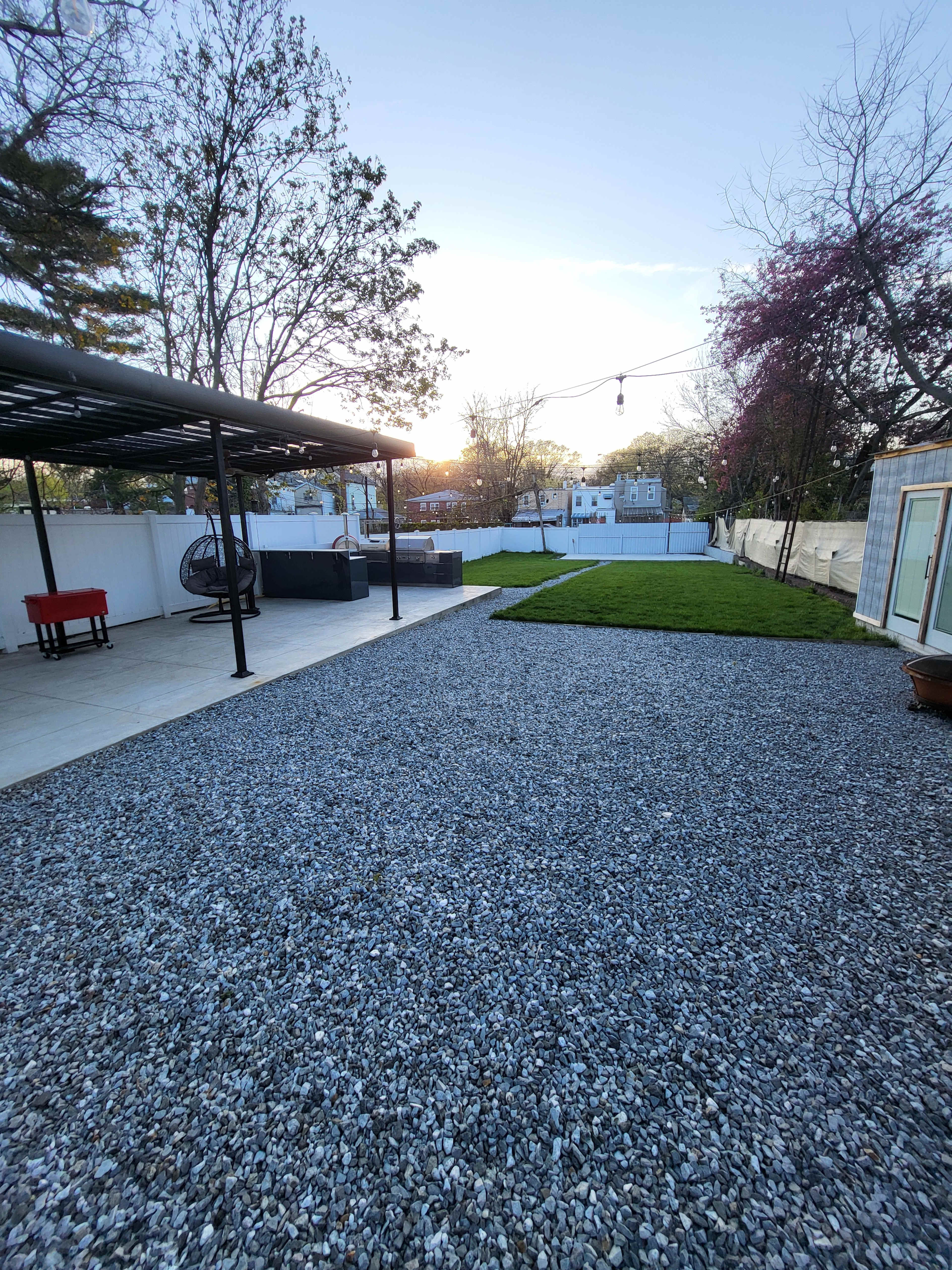 A gravel-covered backyard with a patio area, a green lawn, and a structure equipped with a roof.