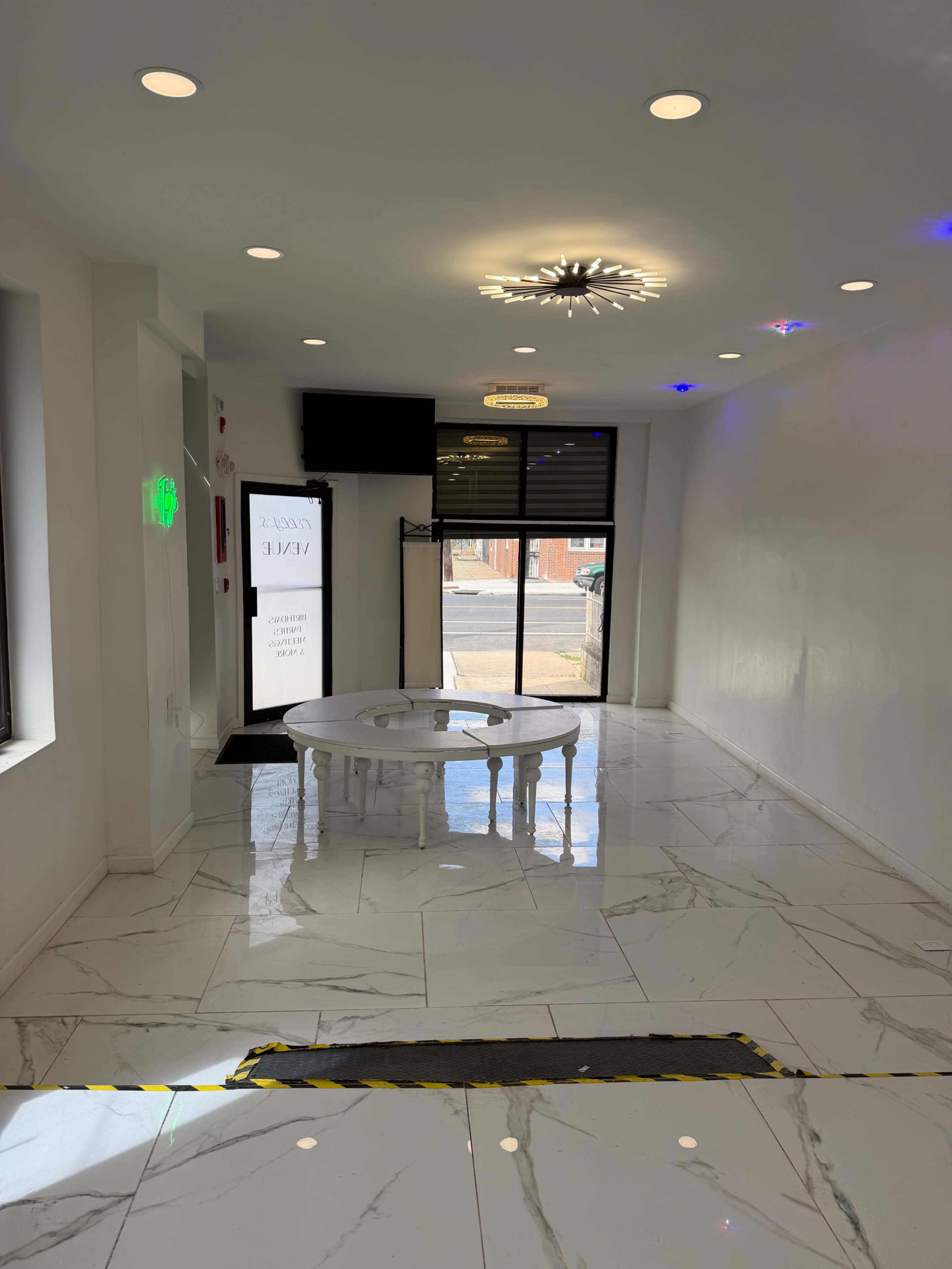 The image shows a modern, minimalist interior of a retail space with a white marble floor, a round table in the center, and a large window at the front.