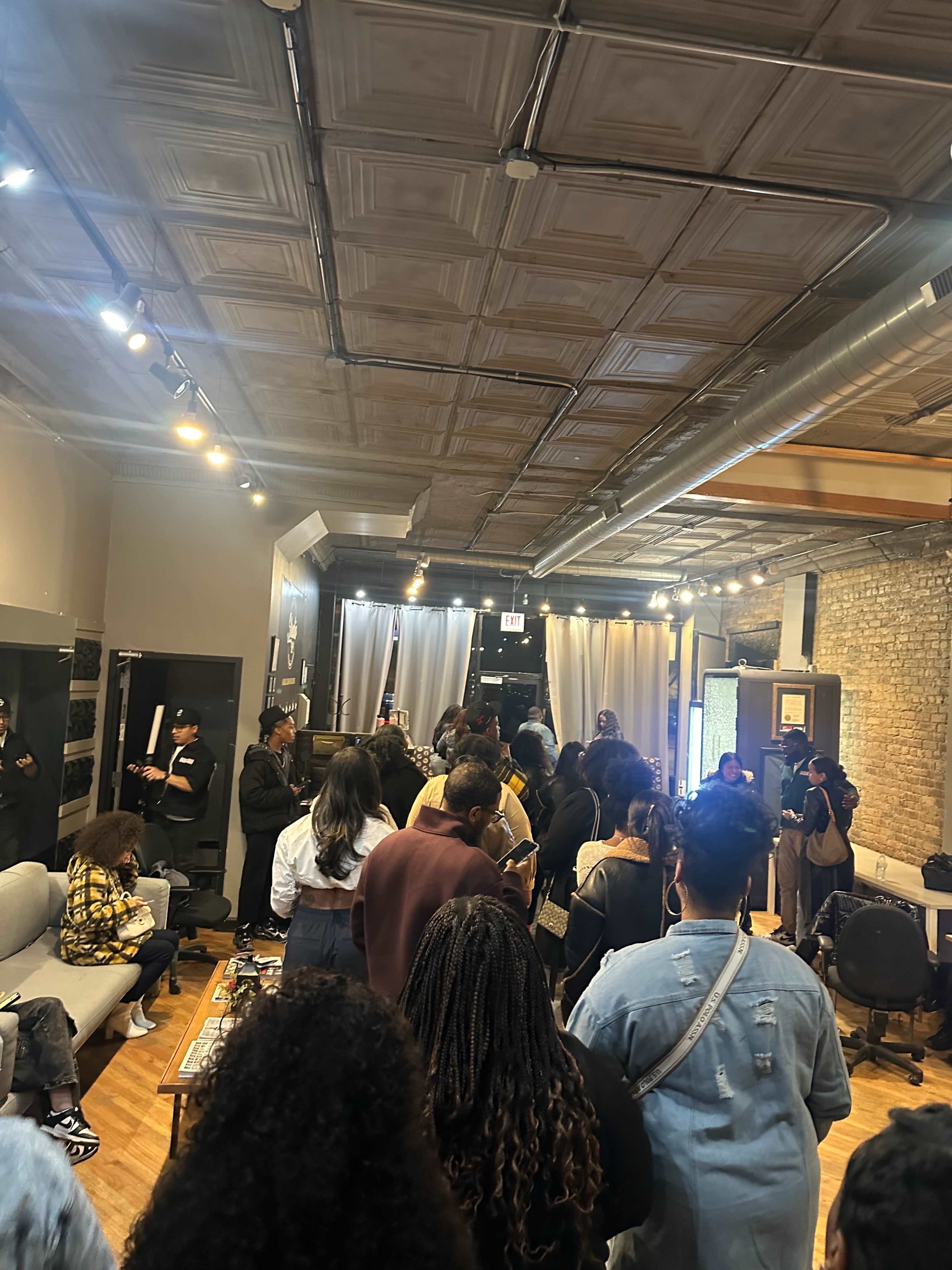 A large group of people stands in a dimly lit room with exposed brick walls, waiting in line while some interact with one another.