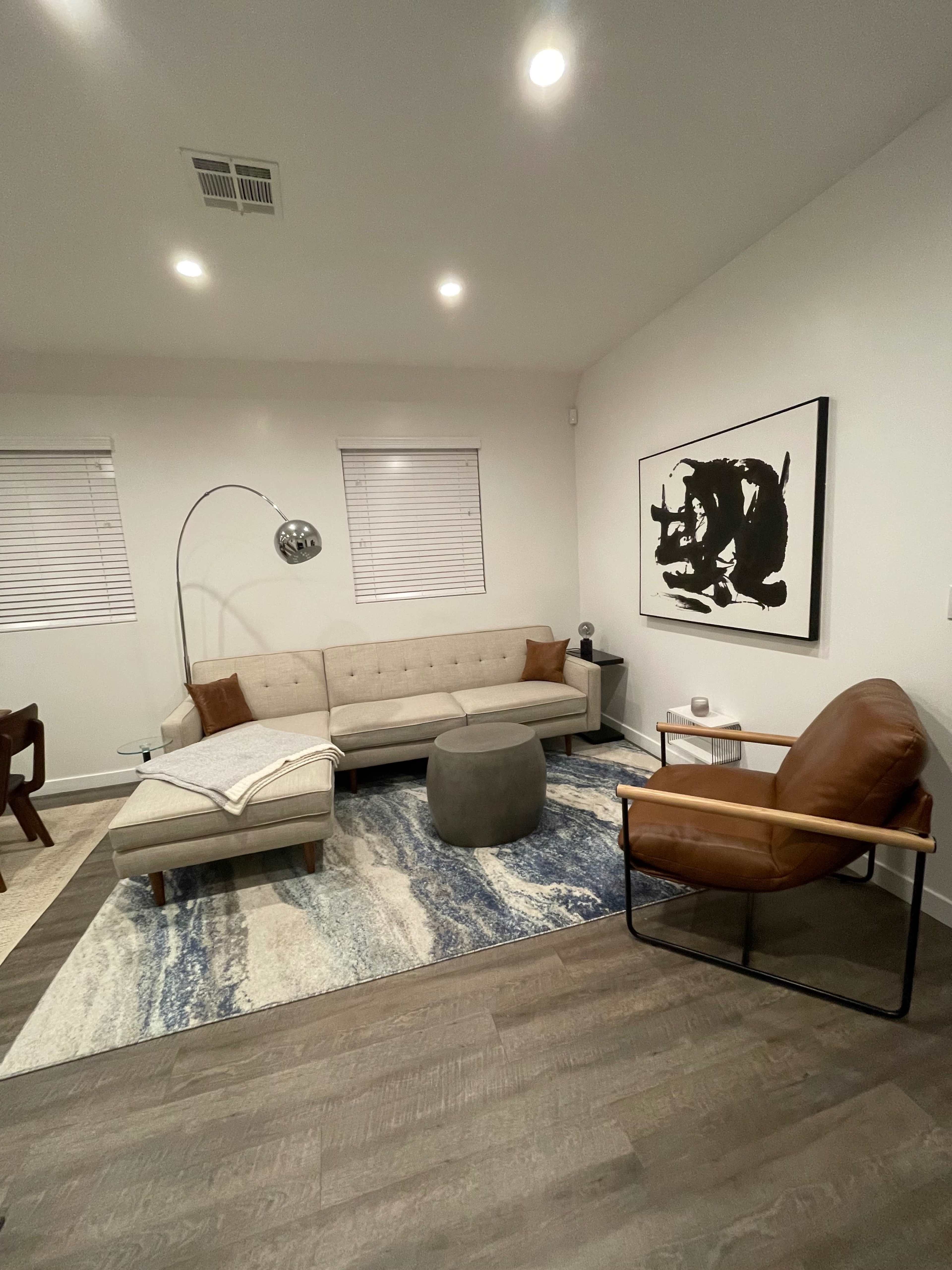 A modern living room features a beige sofa, a brown leather chair, a round ottoman, and an abstract black and white artwork on the wall, all set on a patterned rug.