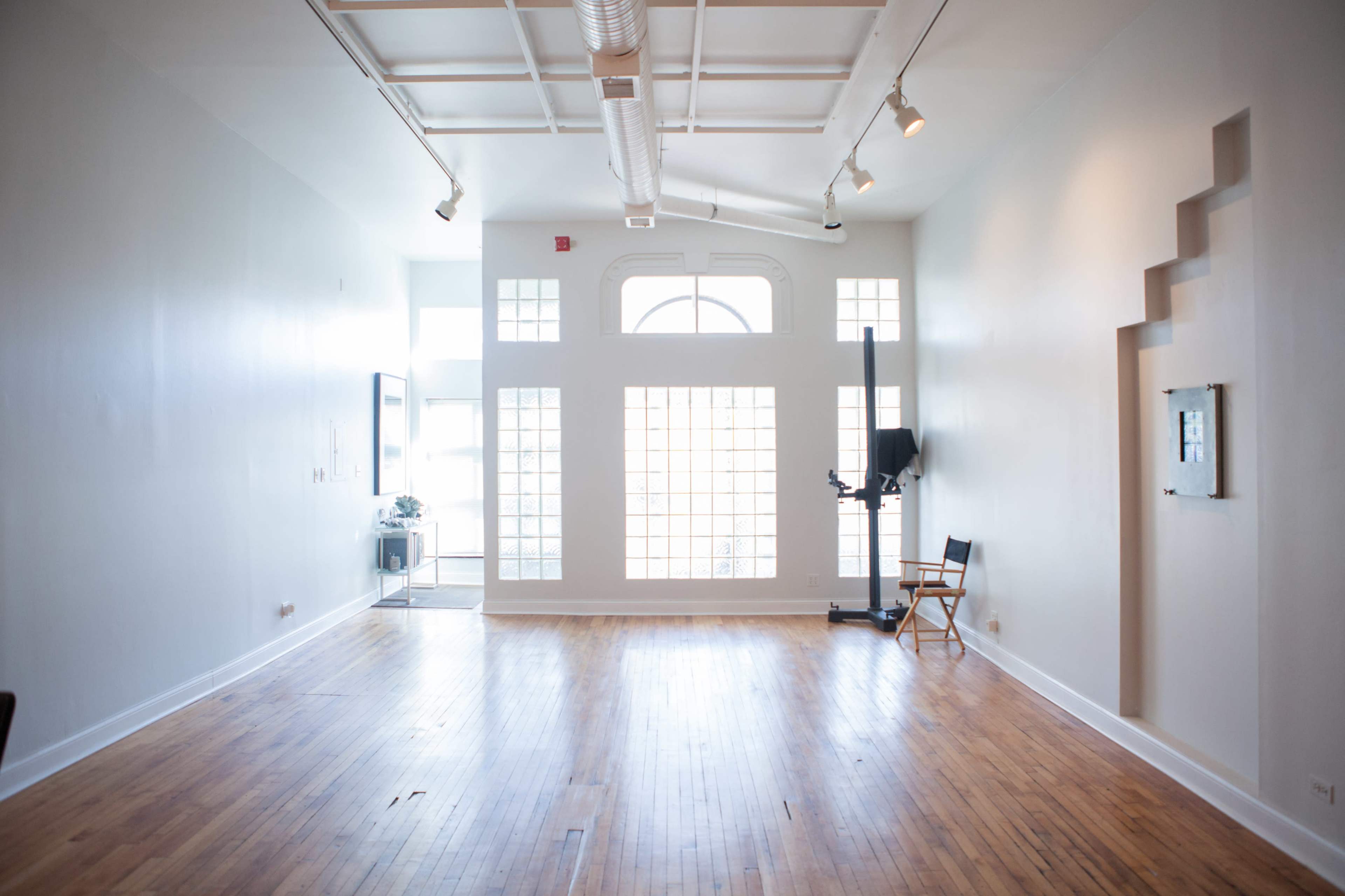 The image shows a spacious, well-lit room with wooden flooring and large windows, featuring minimal furnishings and a single chair.