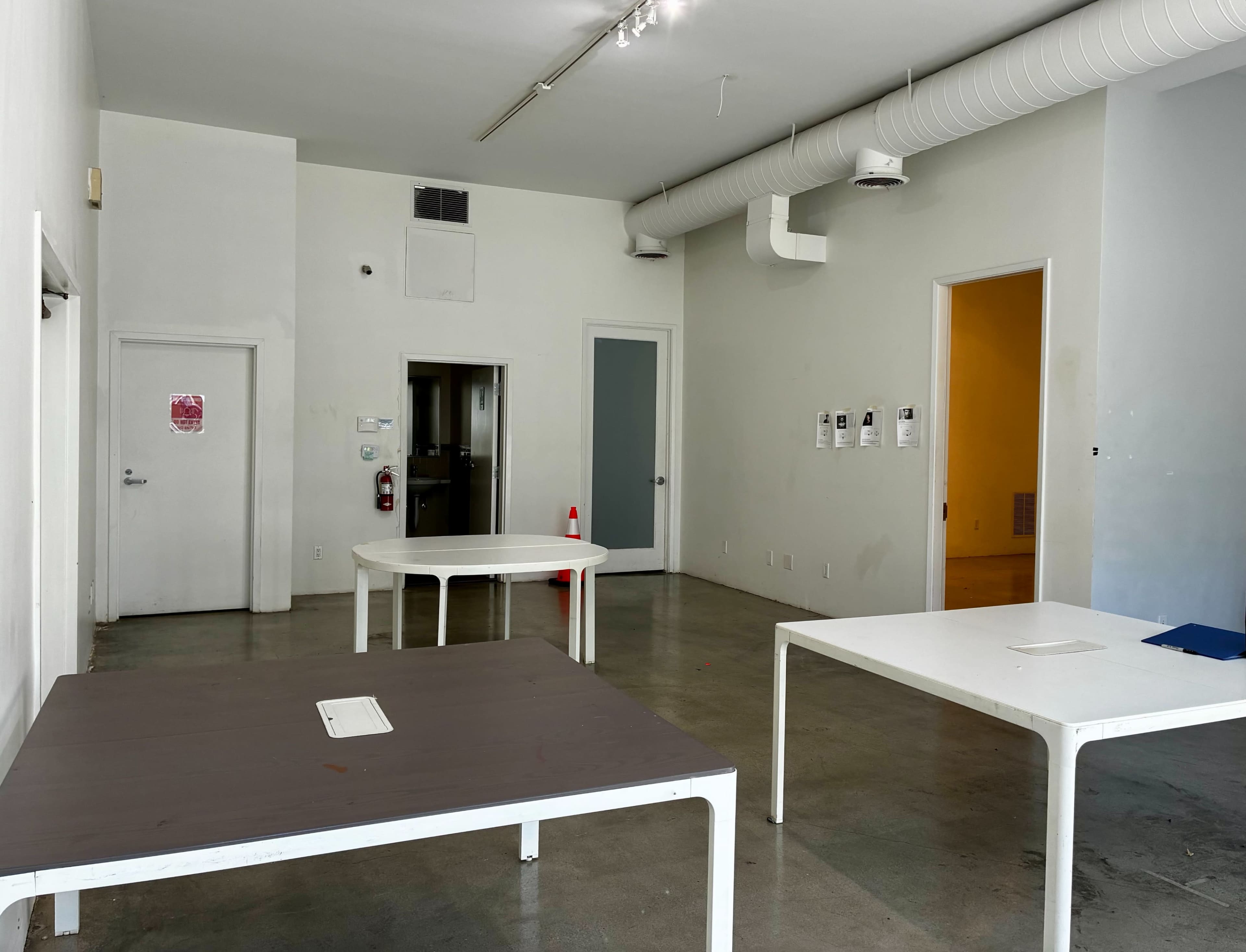 The image shows a sparse room with four white tables positioned around, white walls, and two doors leading to other spaces.
