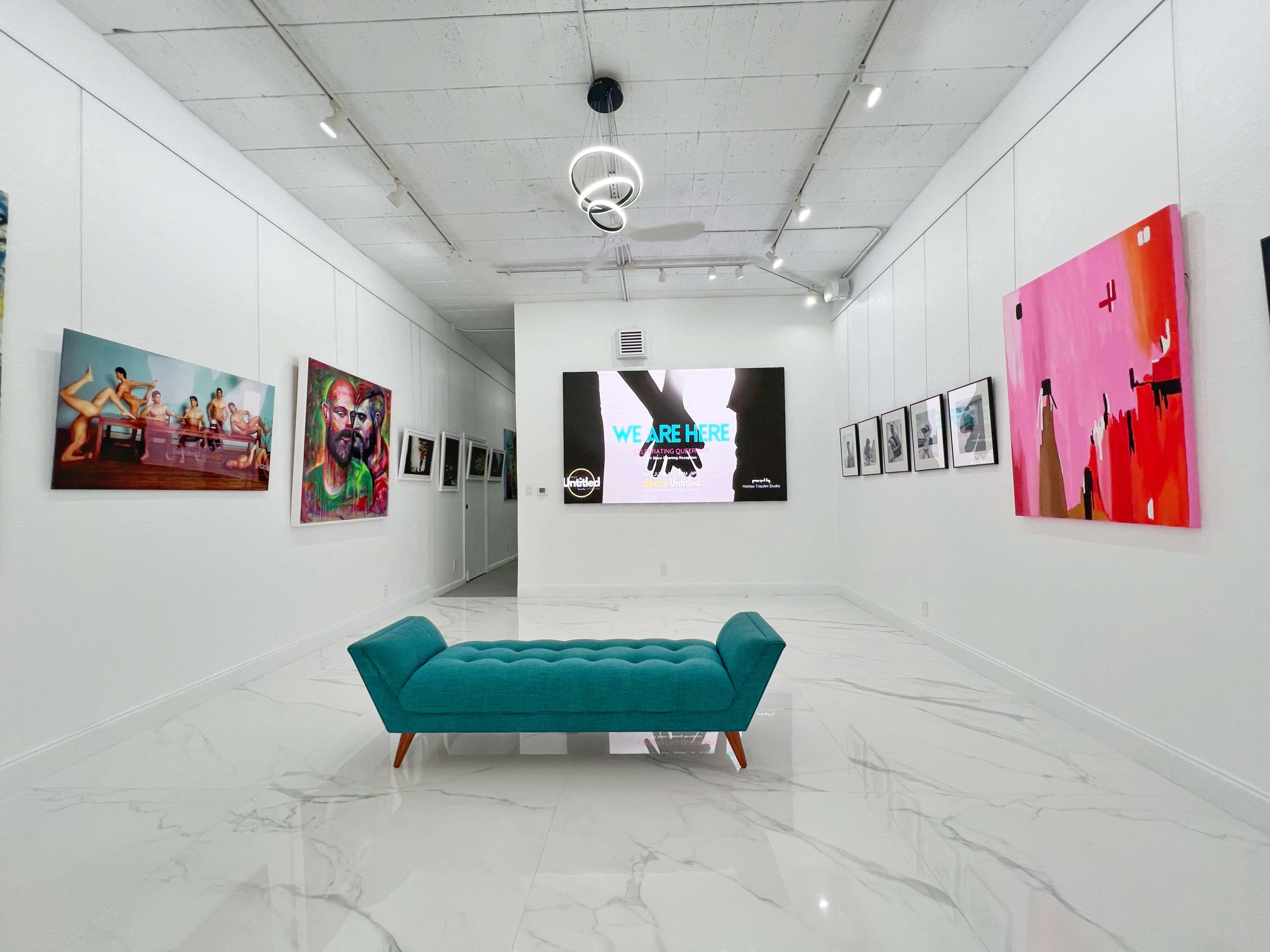 The image shows a modern art gallery featuring diverse artwork on the white walls and a teal couch positioned in the center of the room.