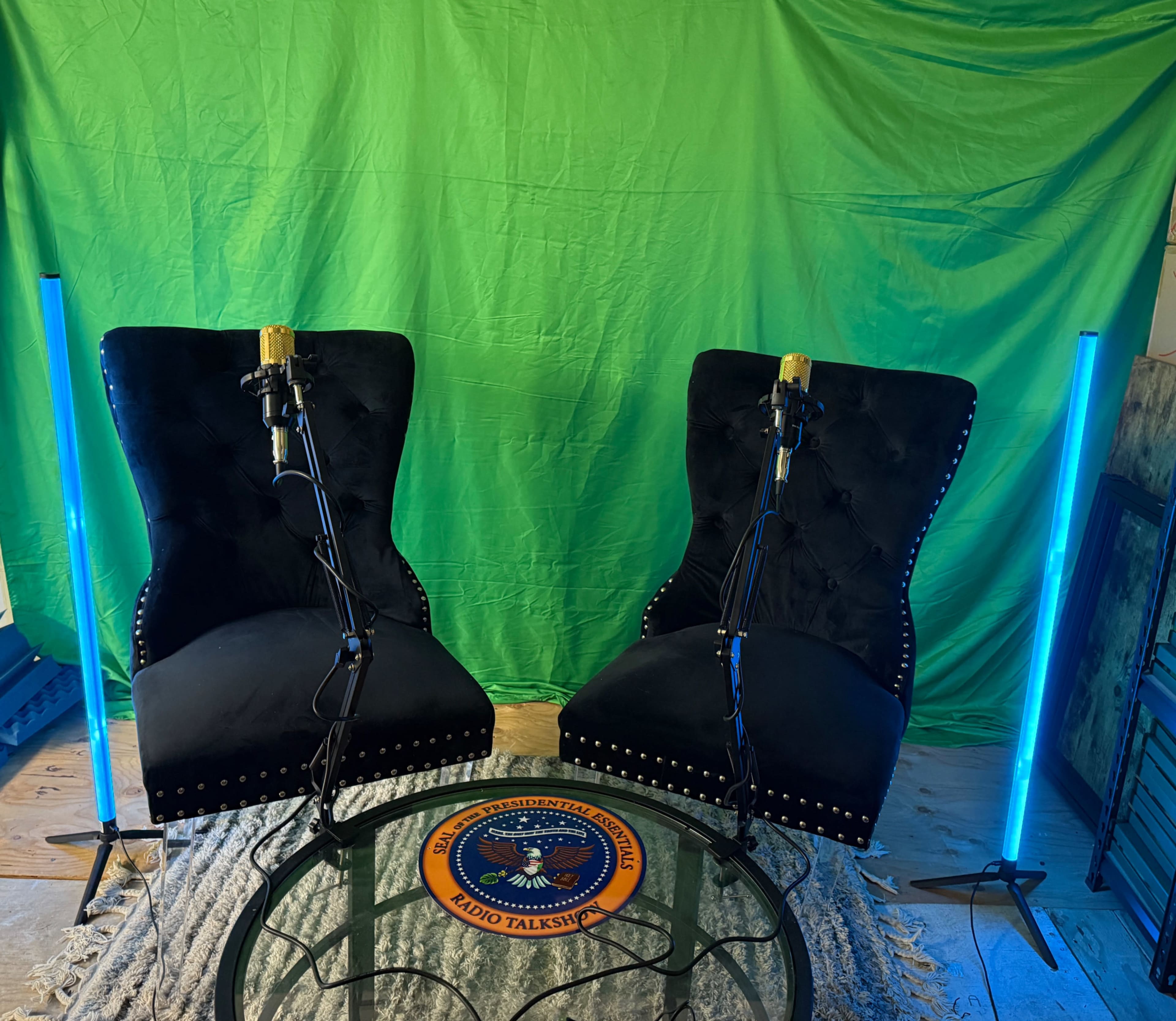 The image shows two black tufted chairs with microphones in front of a green backdrop, accompanied by a round table featuring a logo and illuminated by blue lights.