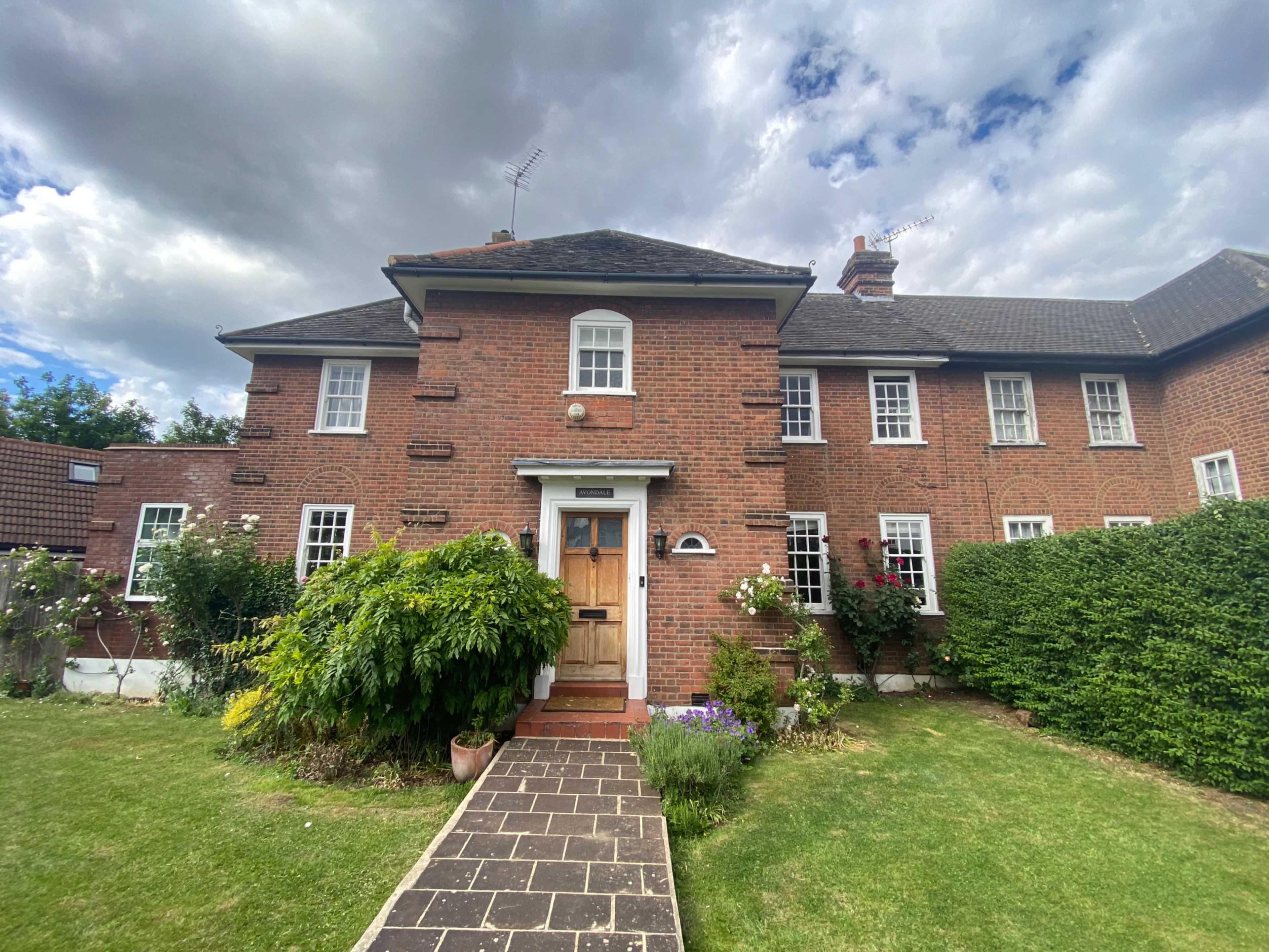 A red-brick house with a symmetrical facade, featuring a central front door surrounded by green shrubs and a well-maintained lawn.