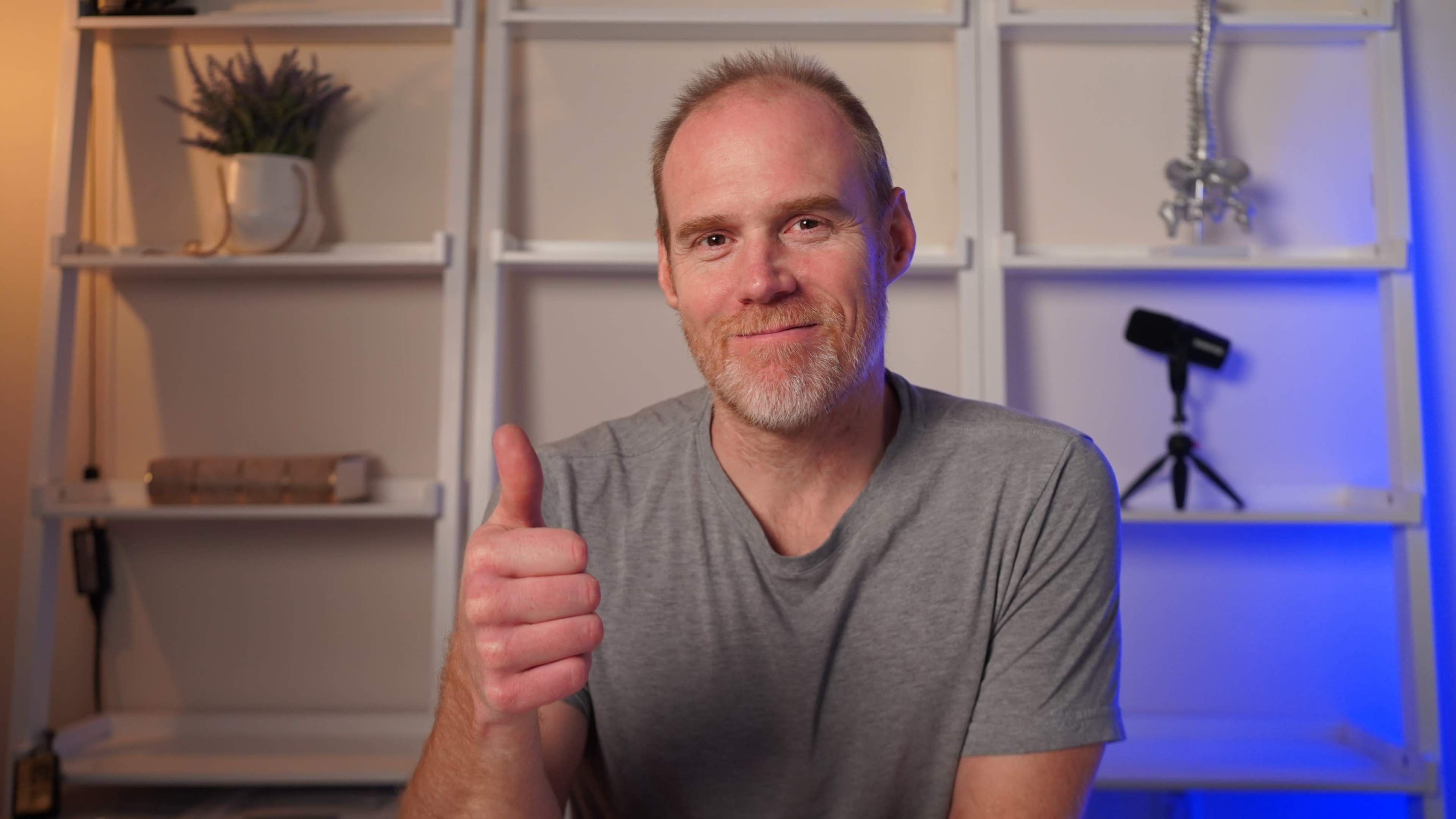 A man in a gray shirt gives a thumbs-up gesture while sitting in front of a shelf with decorative items.