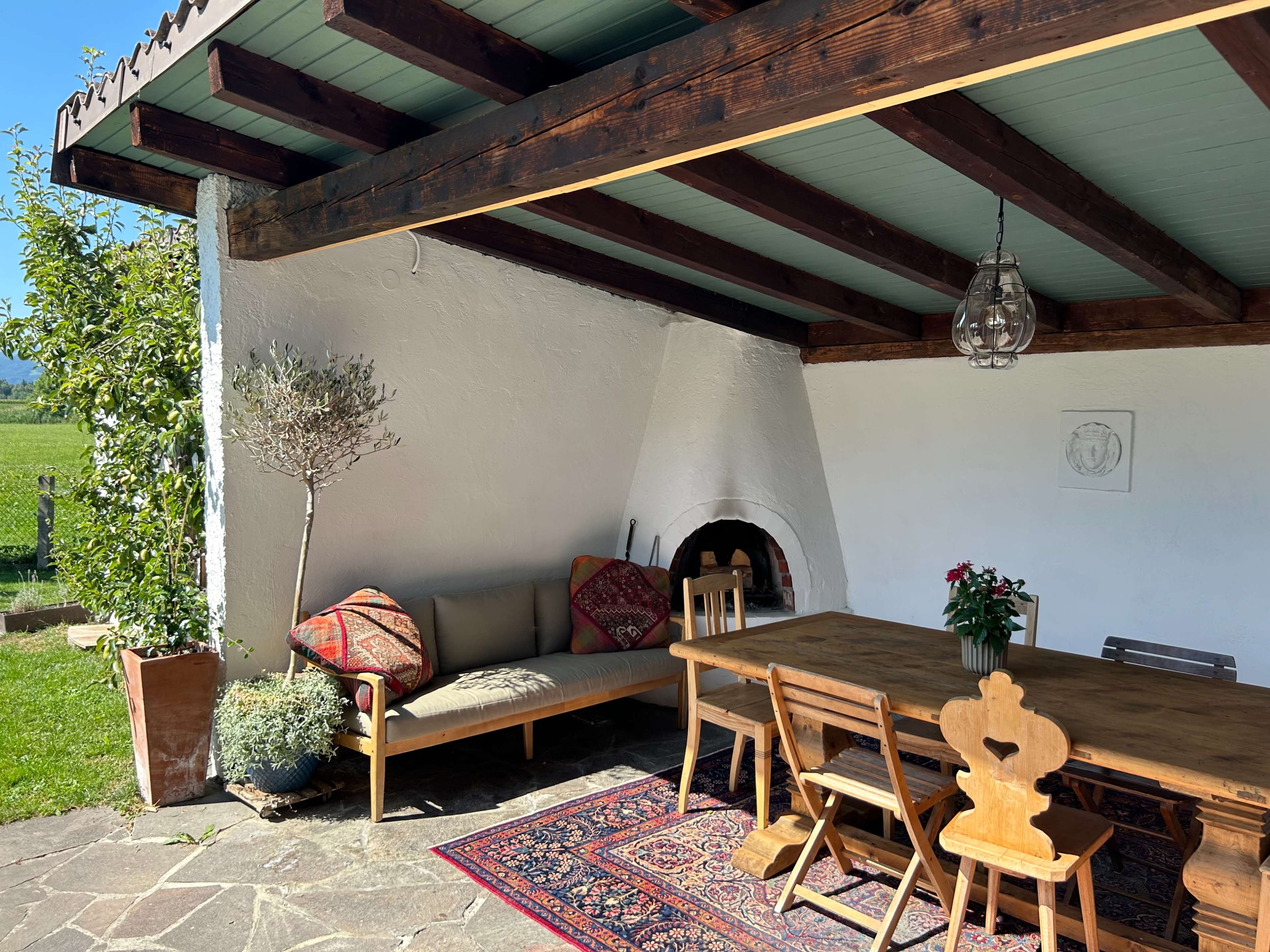 The image shows a spacious outdoor patio with wooden beams, a large dining table, a sofa with decorative pillows, and a potted plant against a white wall.