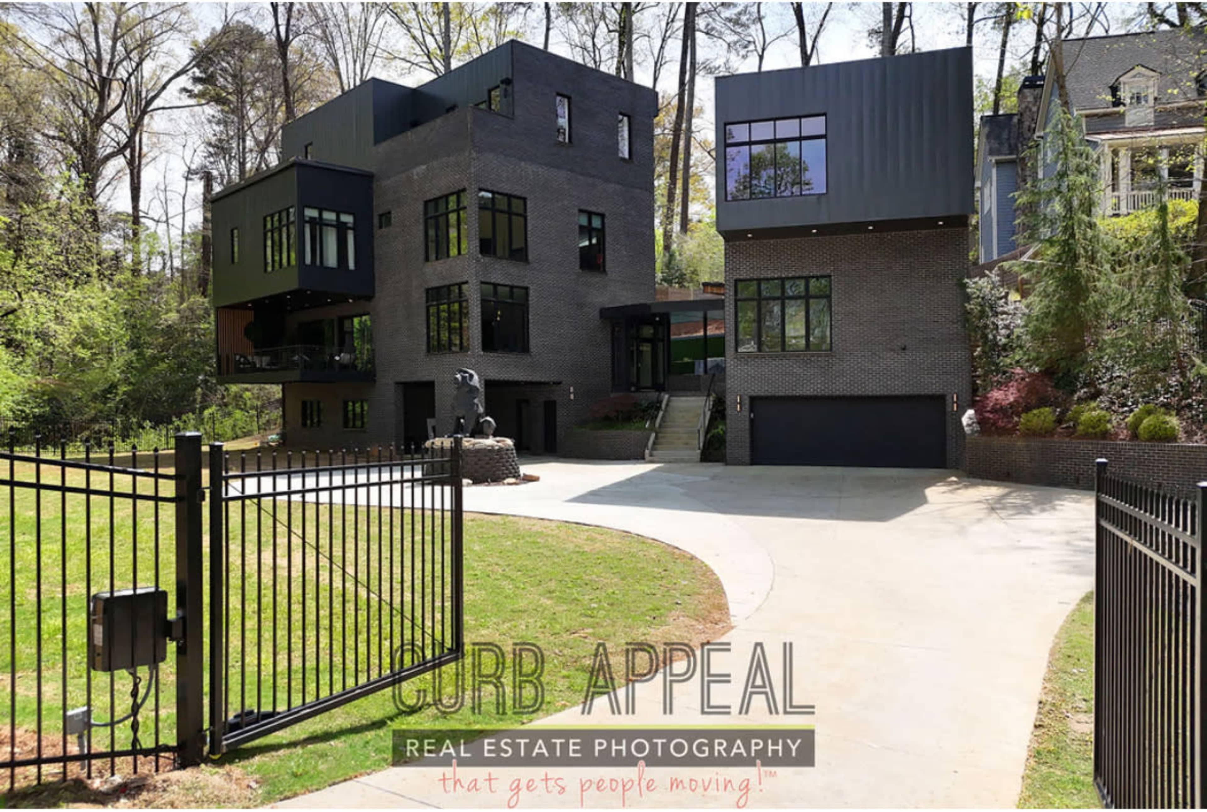 The image shows a modern, multi-level home with a dark facade, large windows, and a circular driveway, set within a wooded area.