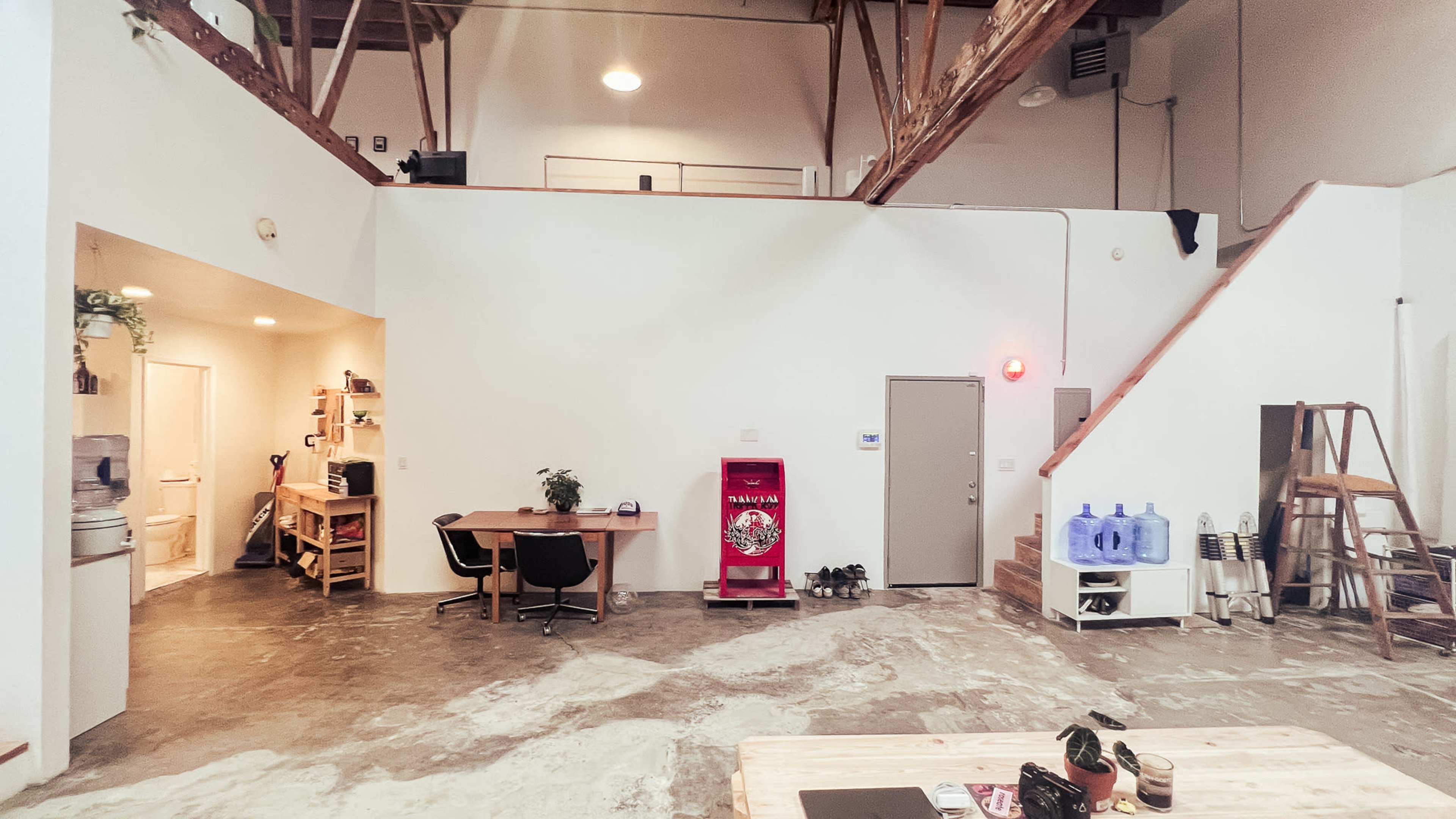 The image shows an open-concept workspace featuring a table with chairs, a water cooler, a red vending machine, and a staircase leading to an upper level.