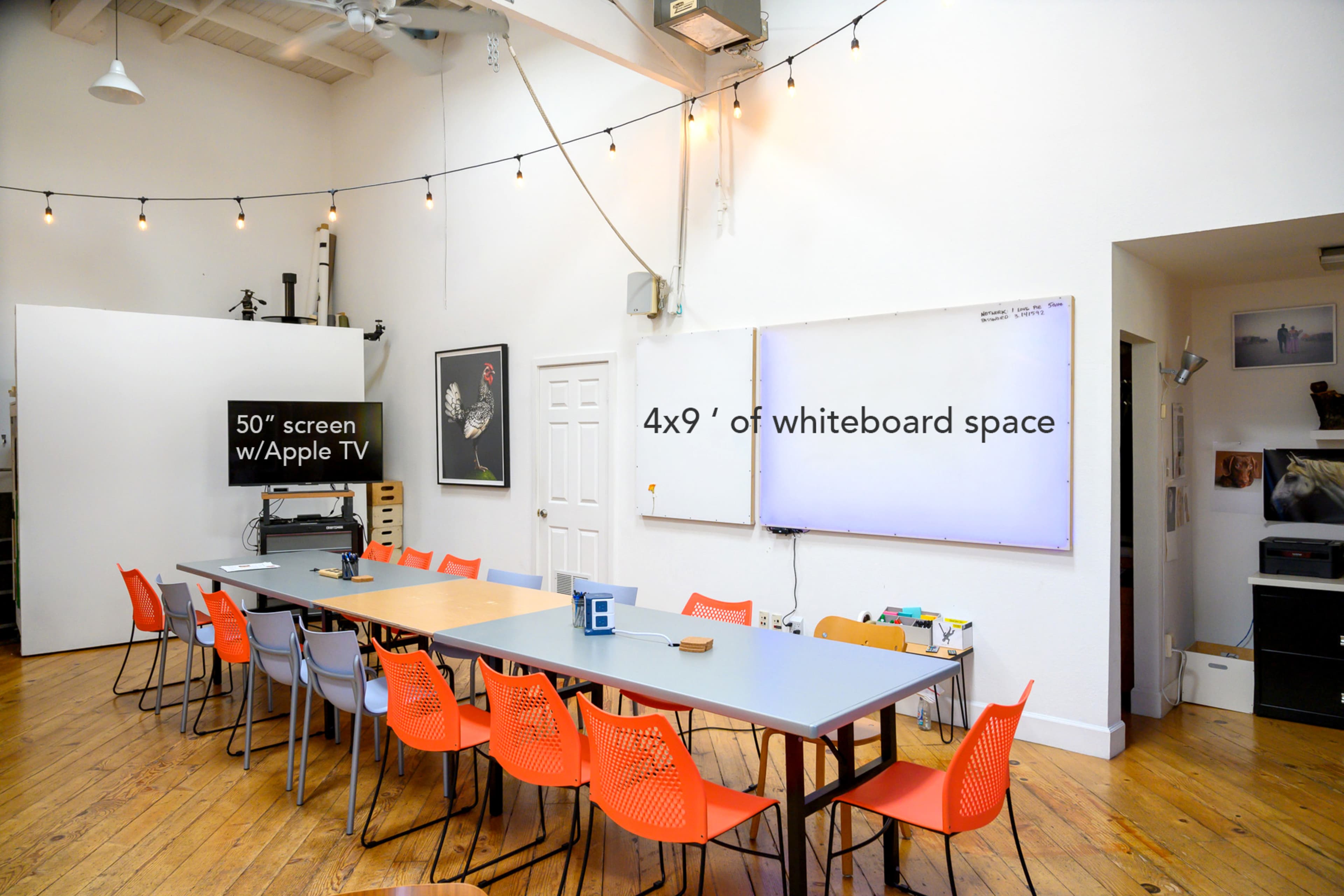 The image shows a spacious meeting room with a long table, orange chairs, a 50-inch screen, and a large whiteboard.