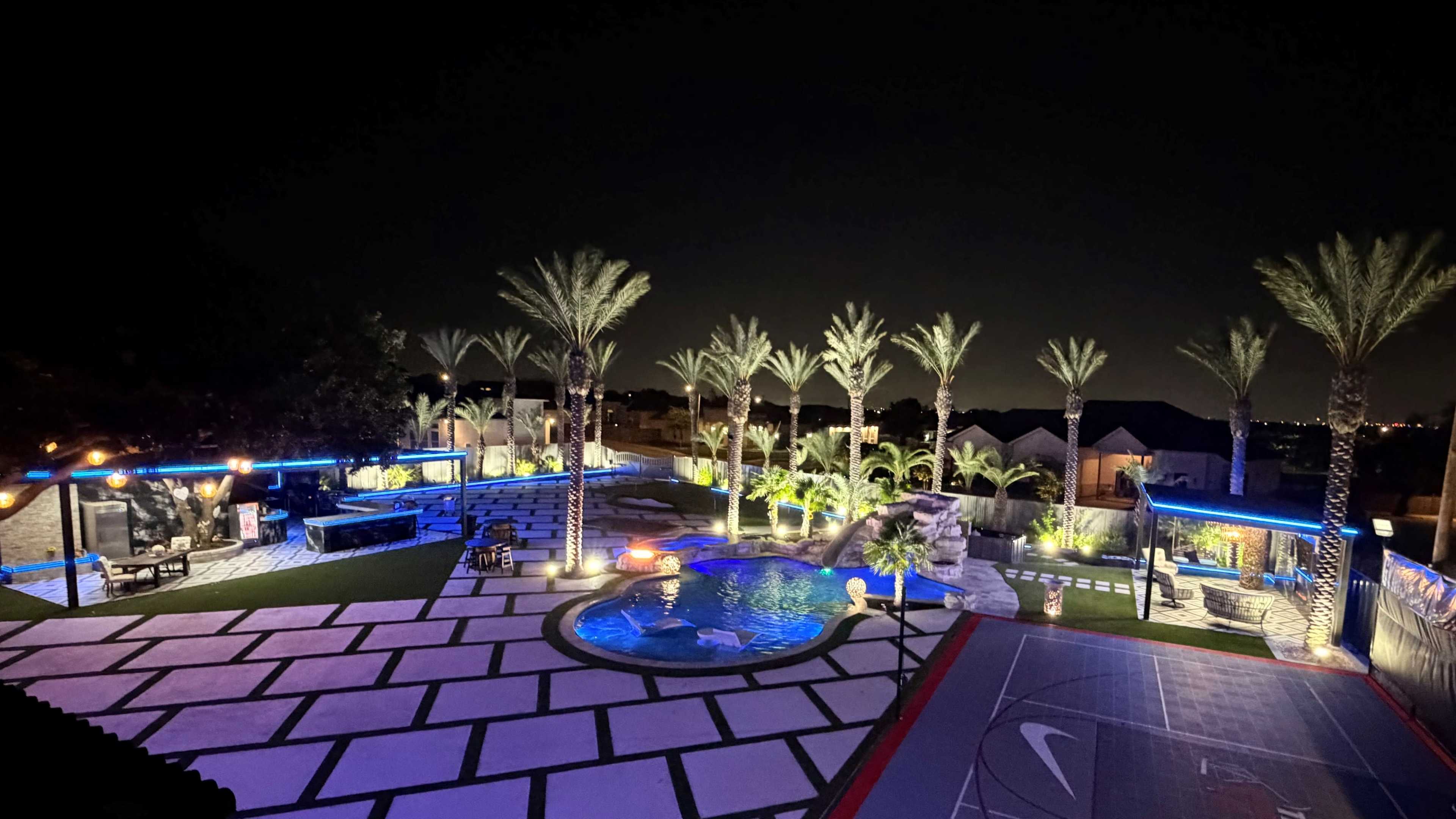 The image shows a well-lit backyard at night featuring a swimming pool, palm trees, a basketball court, and a seating area surrounded by lush landscaping.