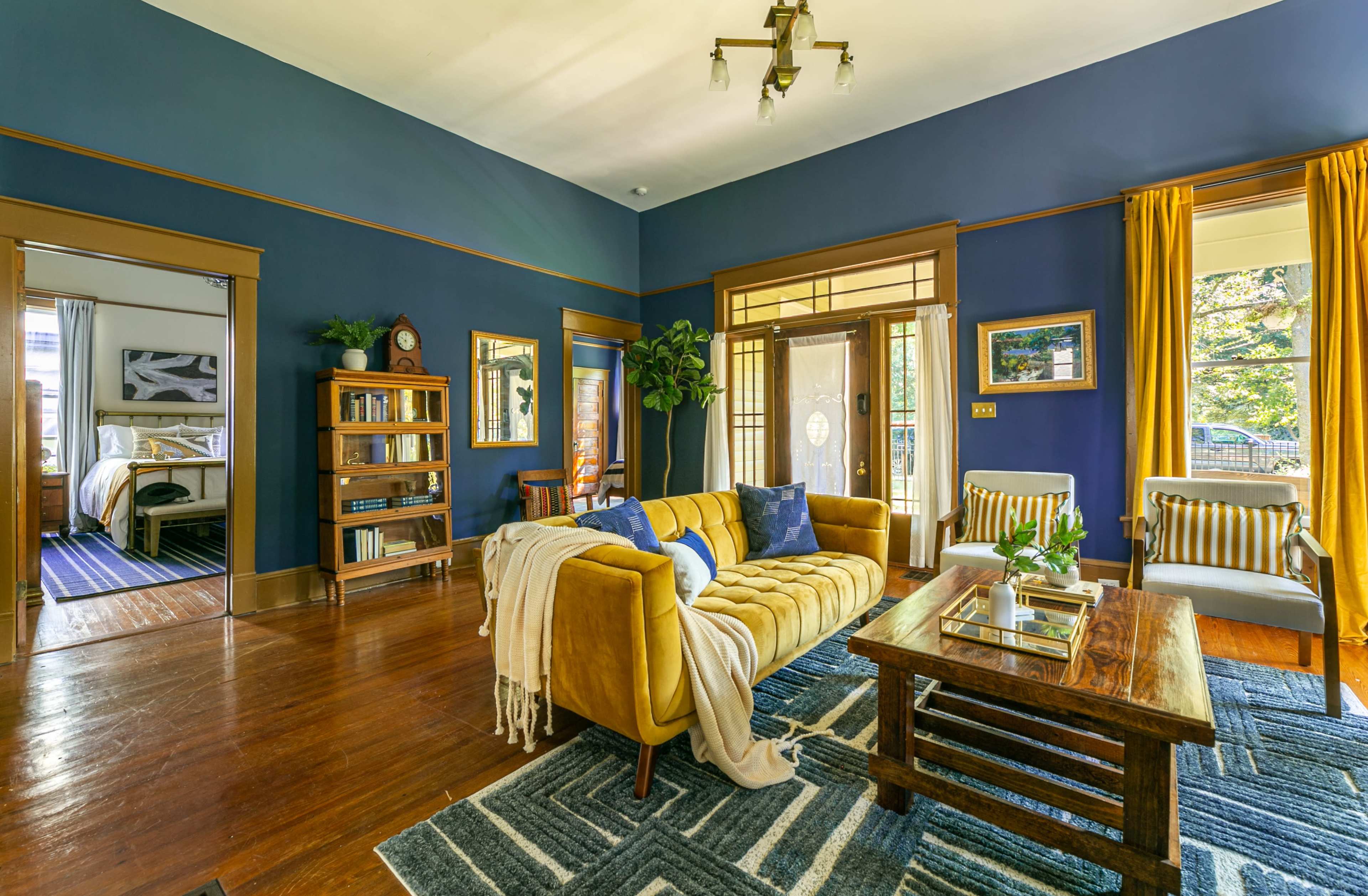 A cozy living room features a yellow sofa, wooden furniture, and blue walls, with a view into an adjoining bedroom.