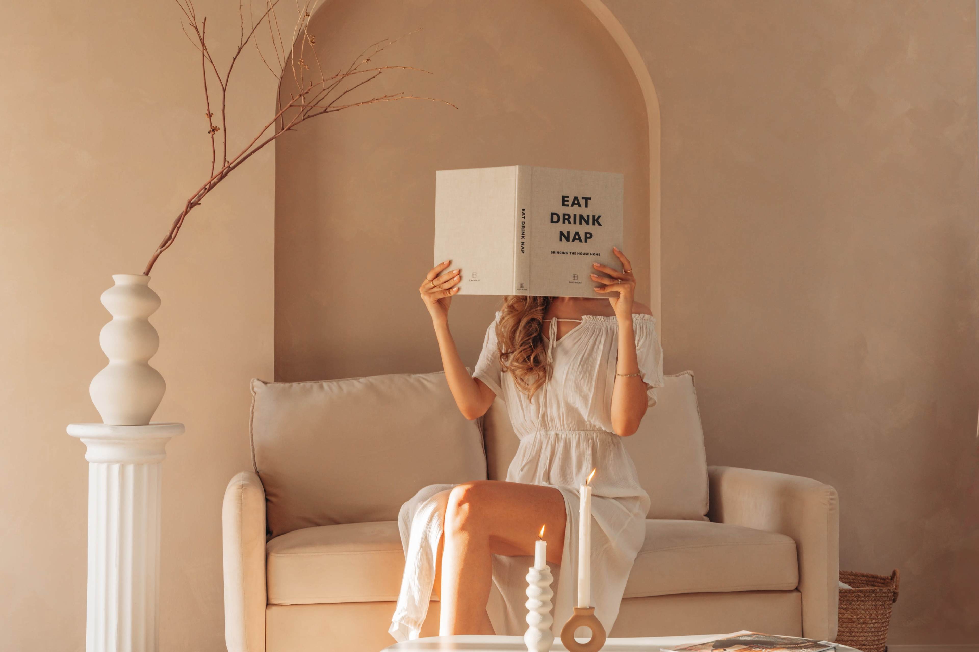 A person sits on a beige sofa, holding a large book titled "EAT DRINK NAP" in front of their face, with a candle and decorative objects on the table.