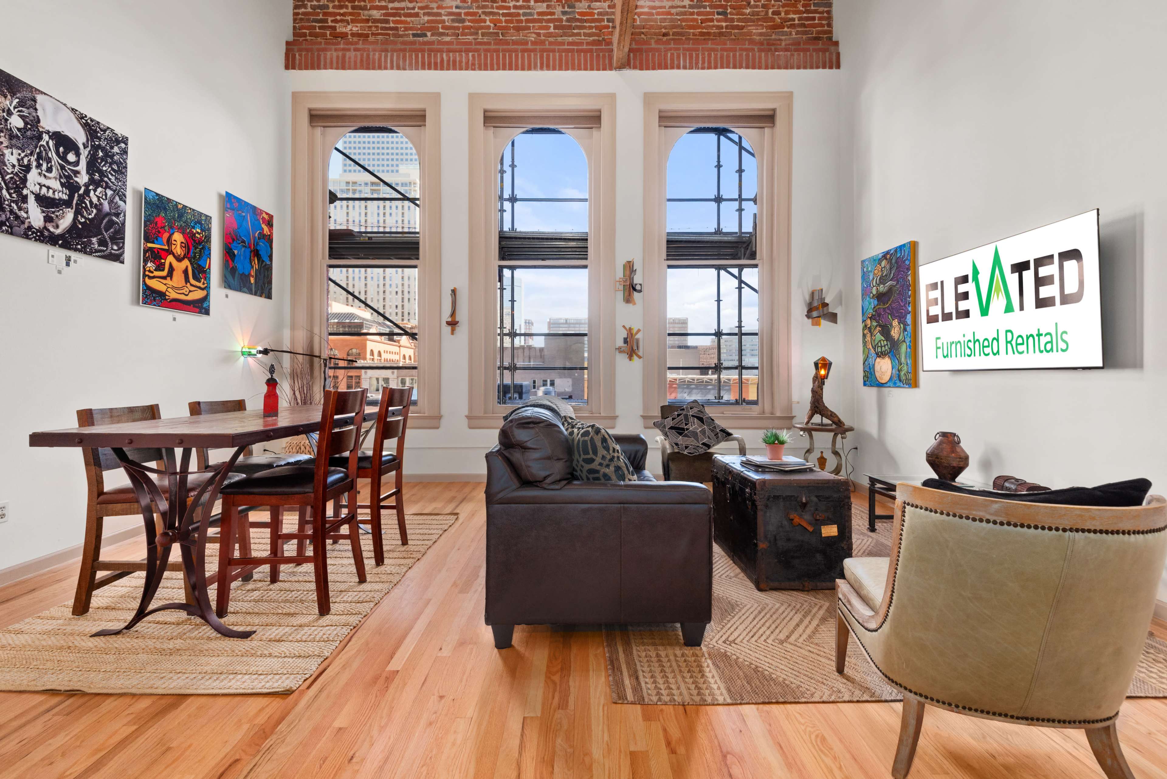 The image shows a modern living space with a dining area featuring a wooden table and chairs, a leather couch, and large windows that allow natural light to enter, highlighting brick walls and various artworks.