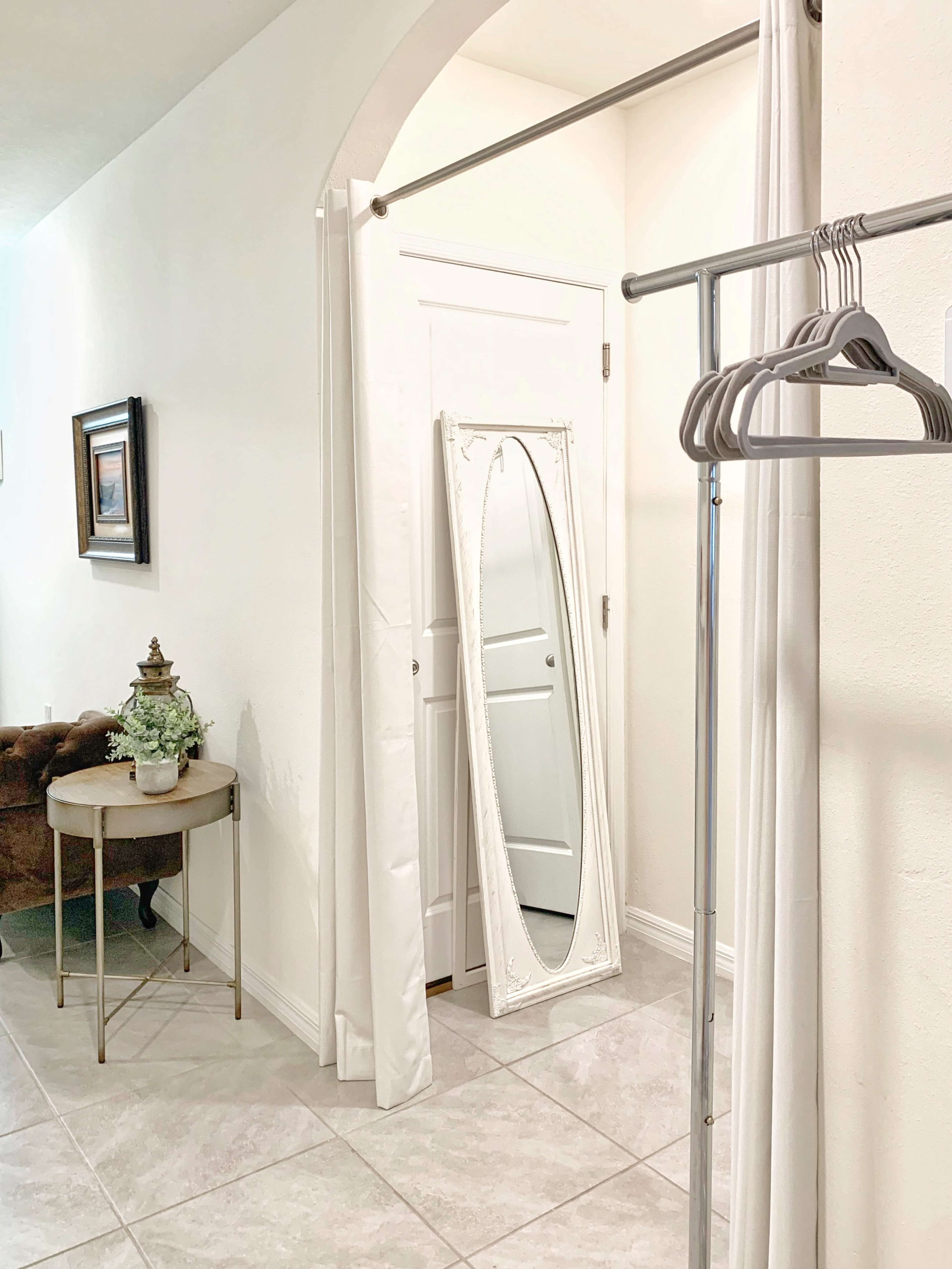 The image shows a hallway with a full-length mirror, a clothing rack with hangers, a small round table, and a brown armchair against the wall.